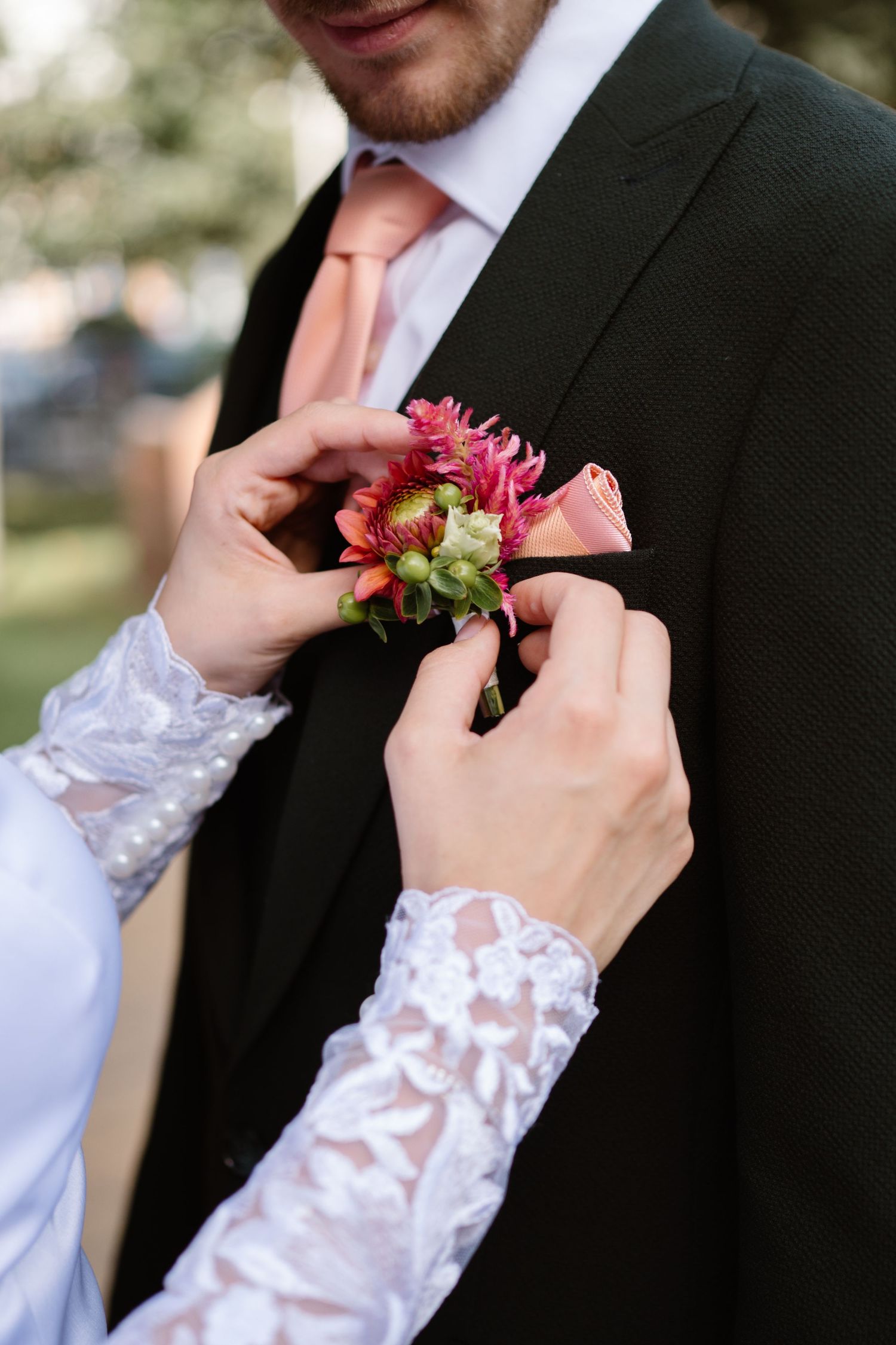 Delicate hands pinning a pink and coral boutonniere onto a dark suit jacket.