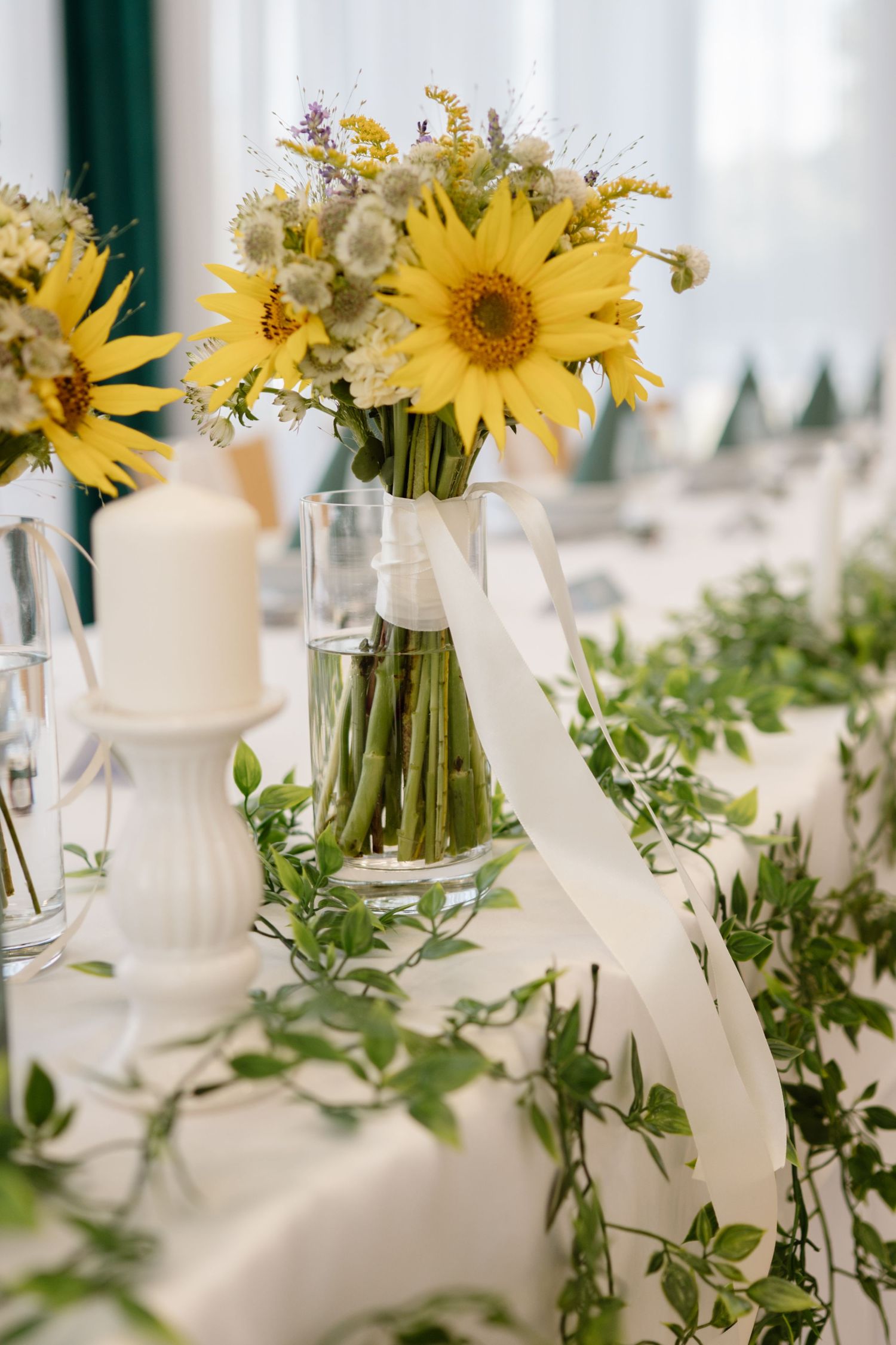Yellow sunflowers and white candles arranged on tables with green foliage.