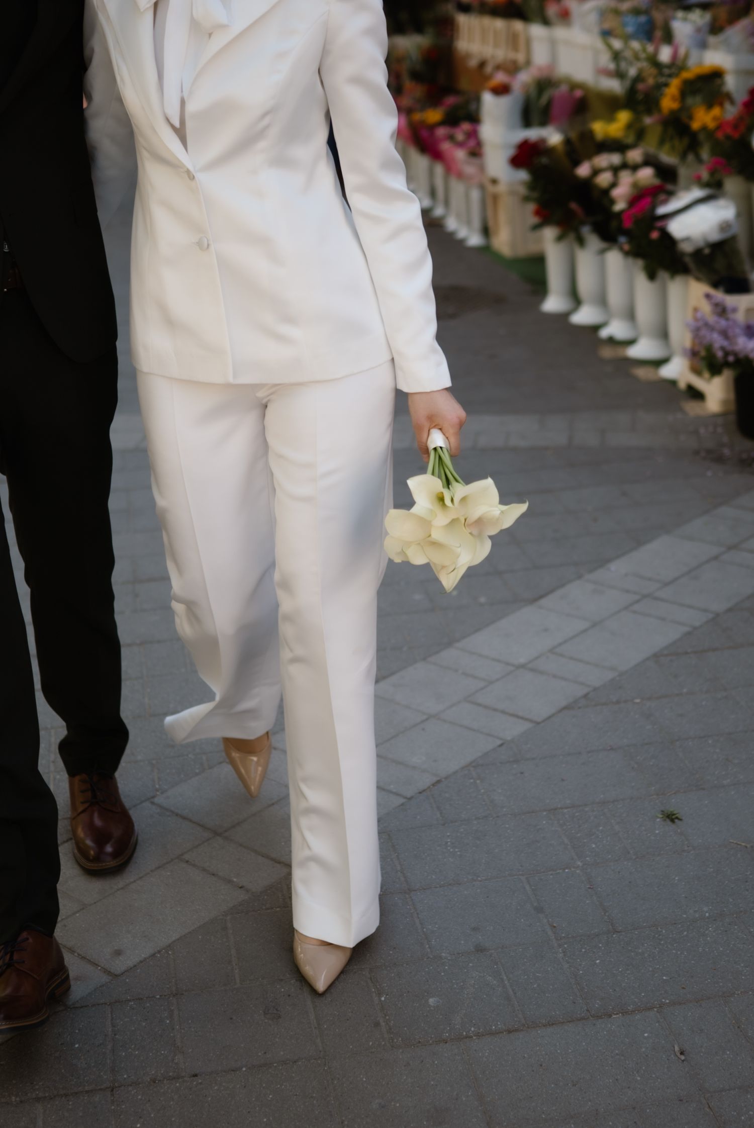 Bride in white pantsuit holding calla lily bouquet walks beside groom on flower market sidewalk.