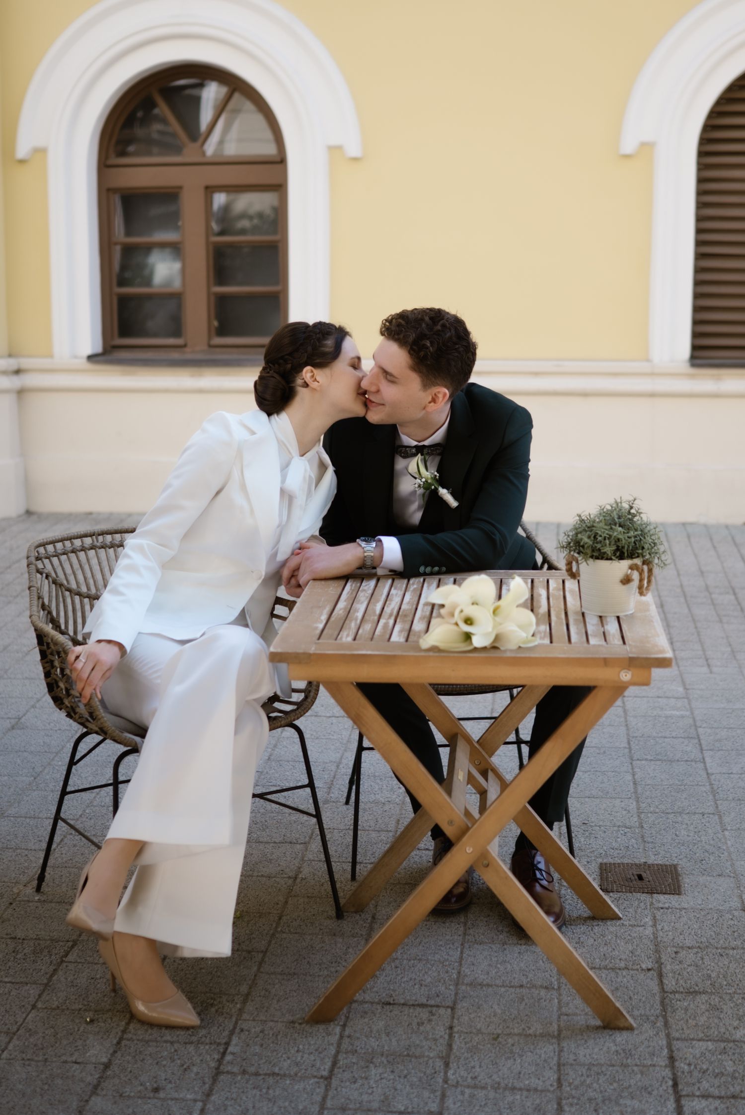 Newlywed couple sharing a kiss at an outdoor wooden table, bride in white suit and groom in black tuxedo.