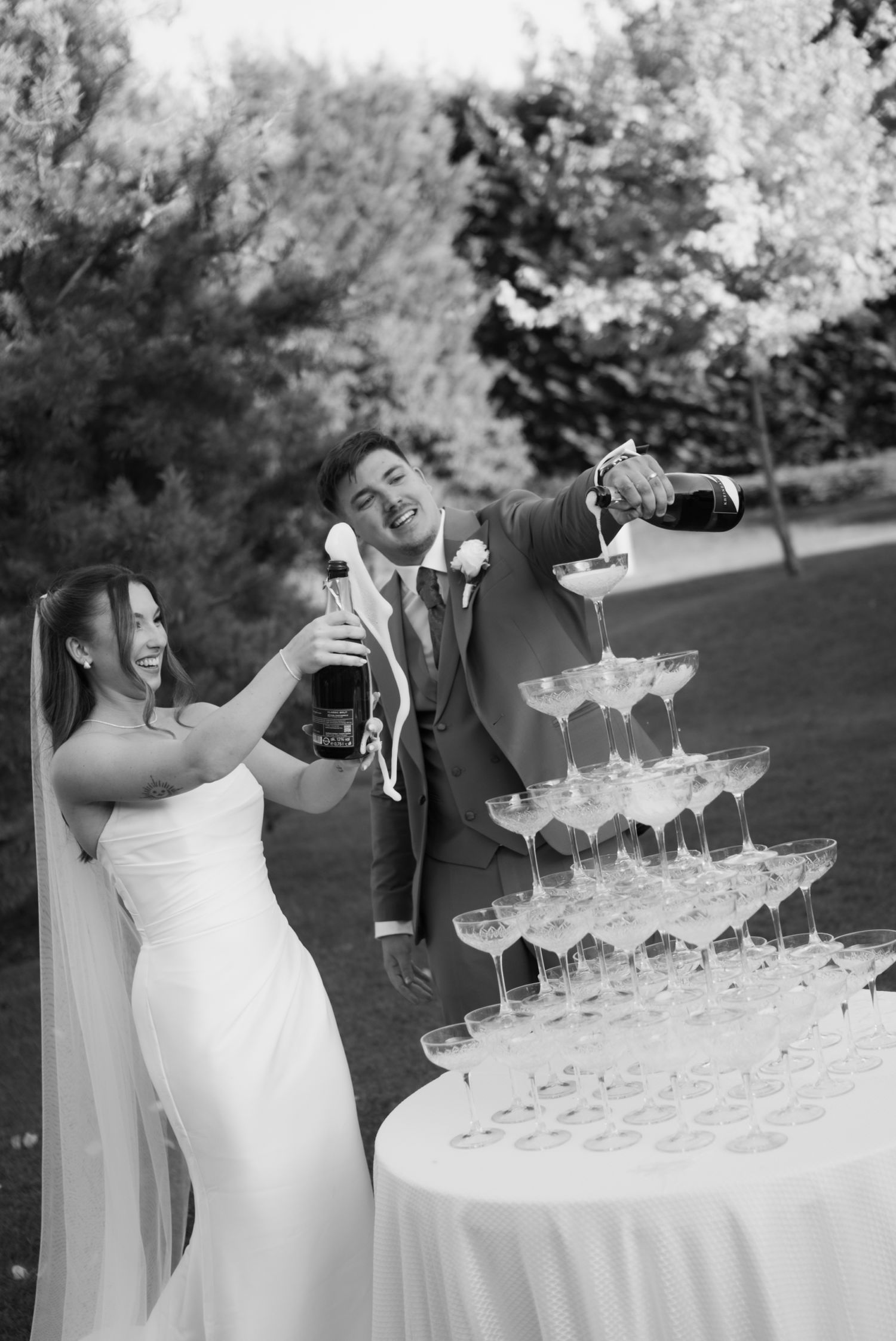 Bride and groom laughing while pouring champagne tower at outdoor wedding reception in black and white.