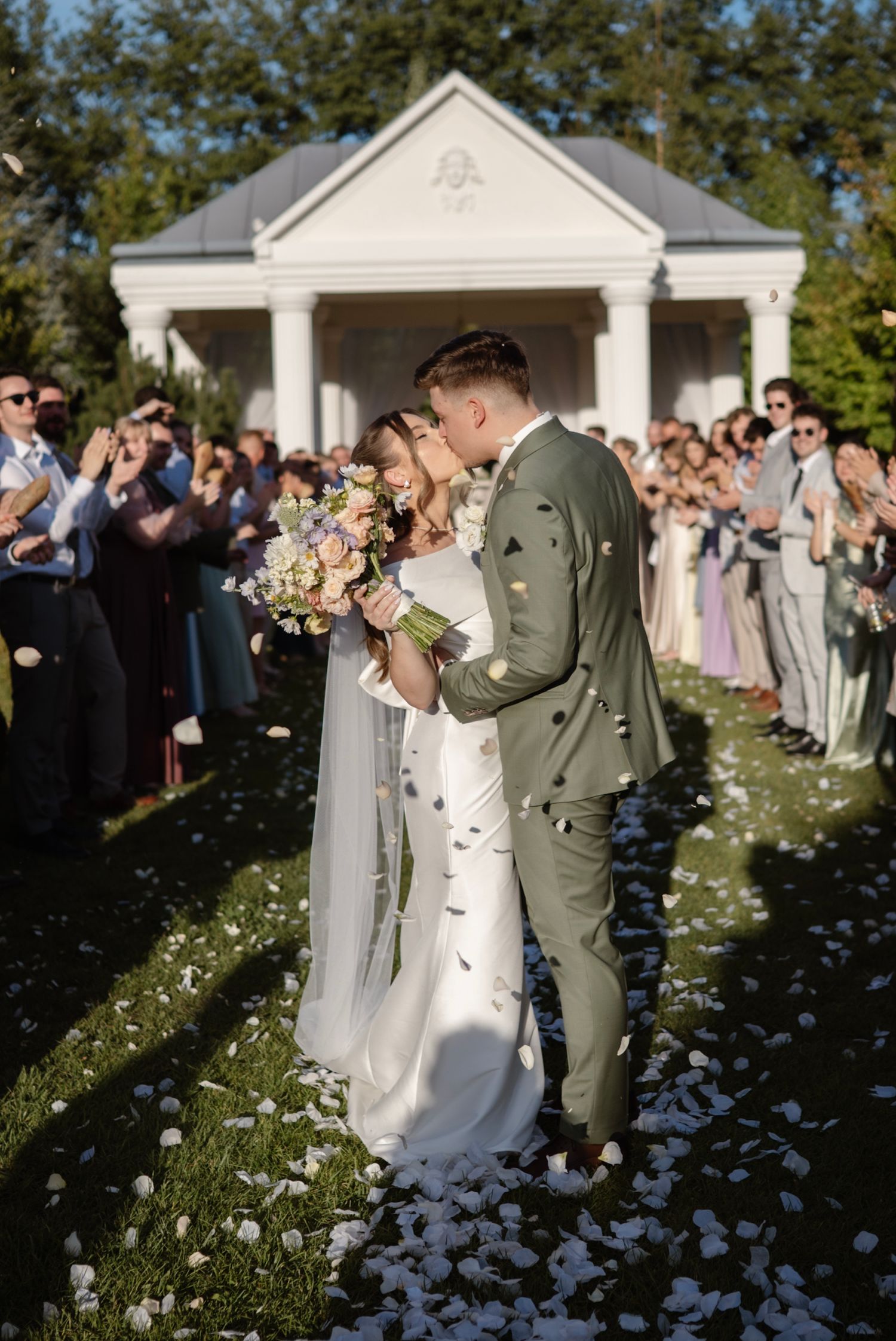 Newlyweds share a kiss while guests toss white flower petals outside a white chapel wedding venue.