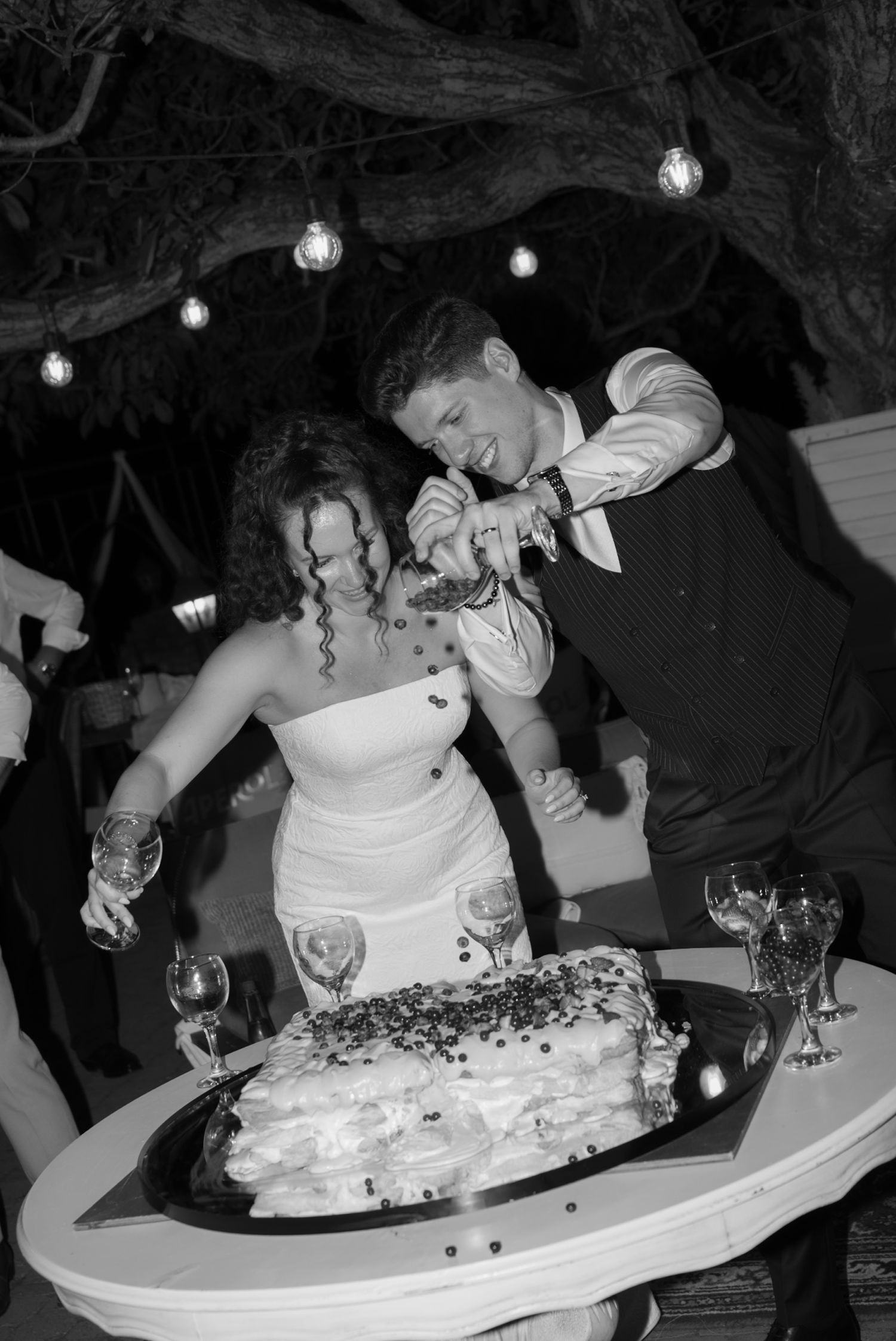 Bride and groom cutting wedding cake together under string lights in black and white photo.