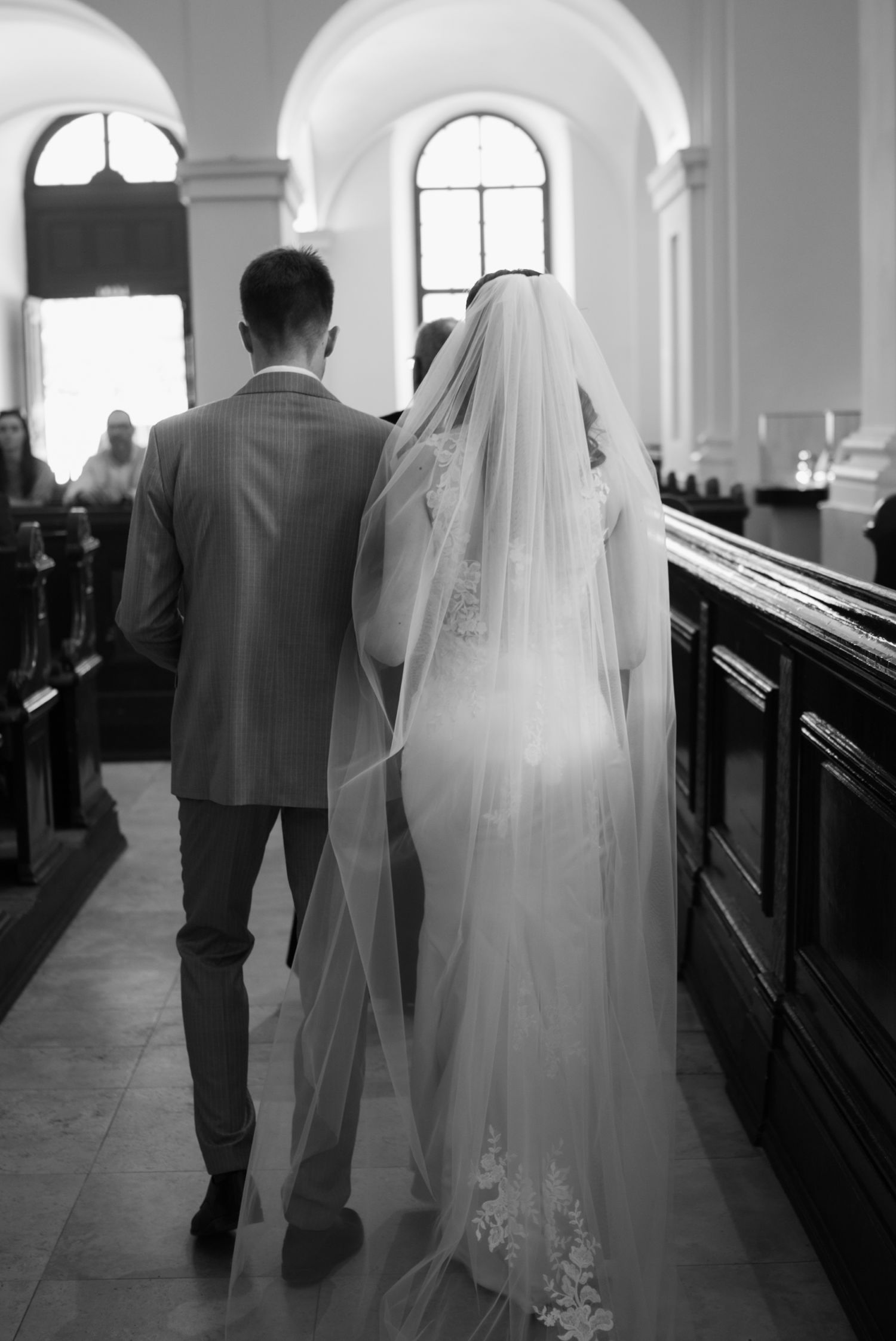 Black and white photo of bride and groom walking down church aisle, bride wearing long veil and lace gown.