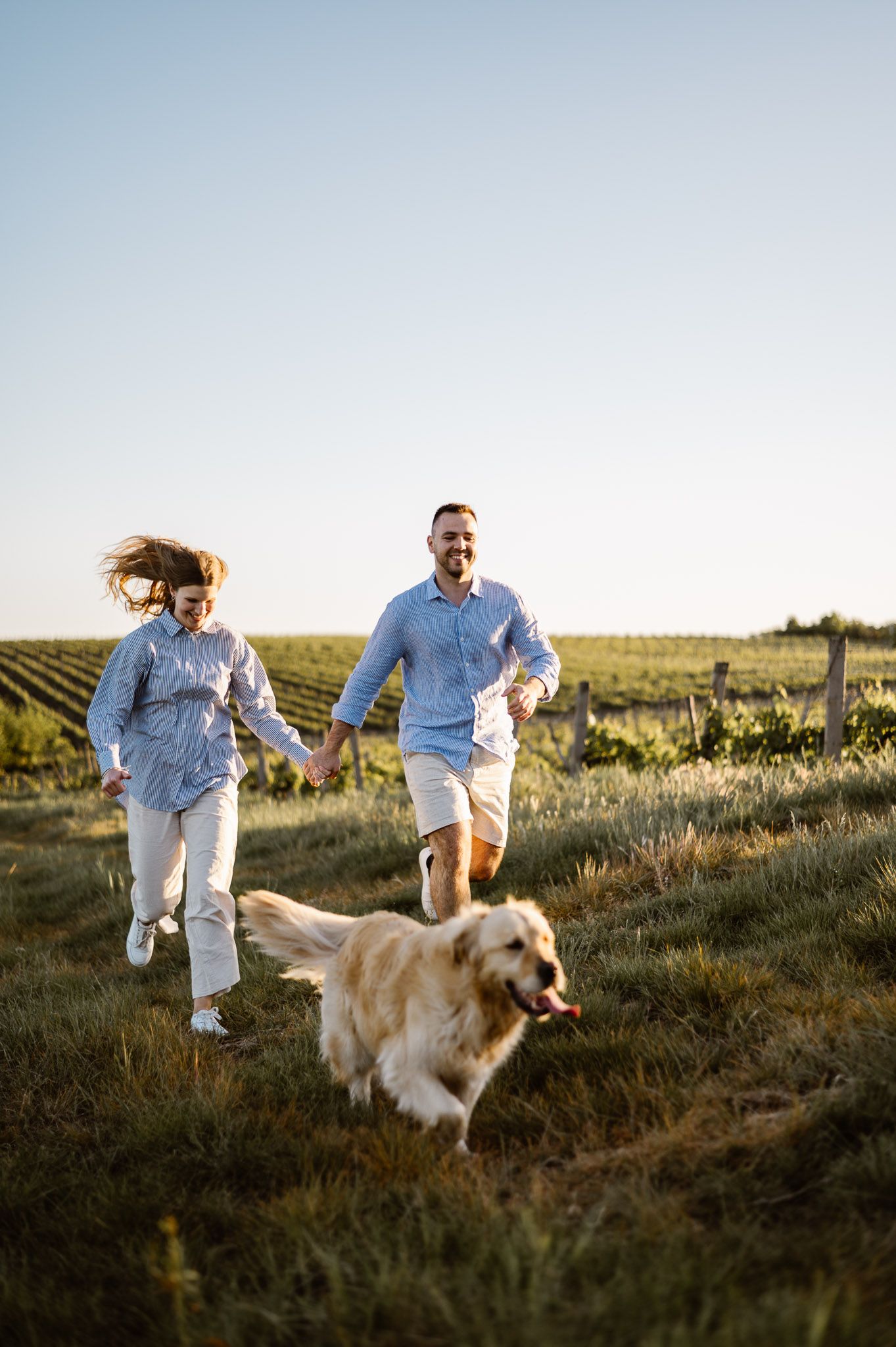 Happy couple running through a field with their golden retriever during golden hour.