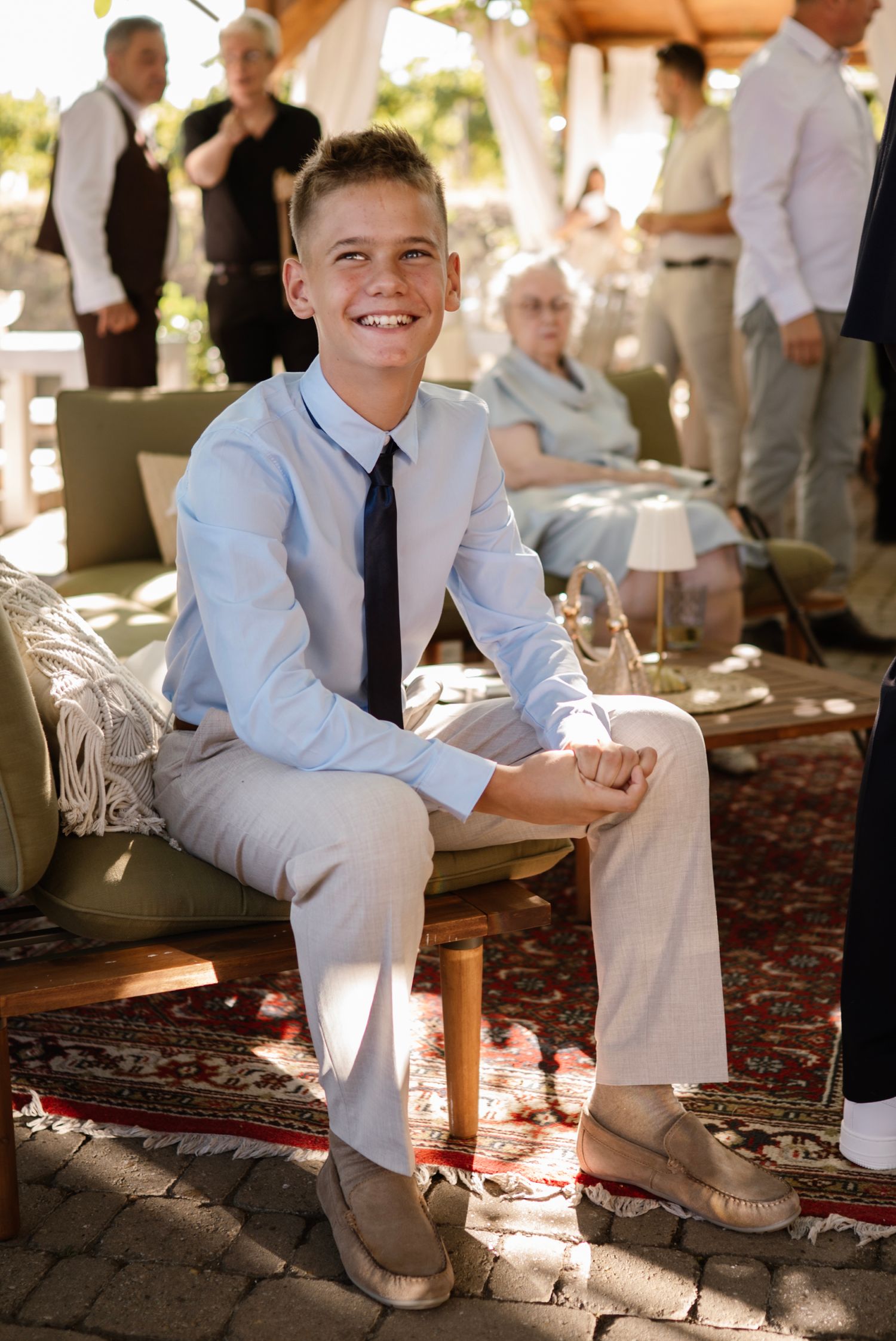 Smiling boy in light blue shirt and navy tie sits at an outdoor wedding reception on a sunny day.
