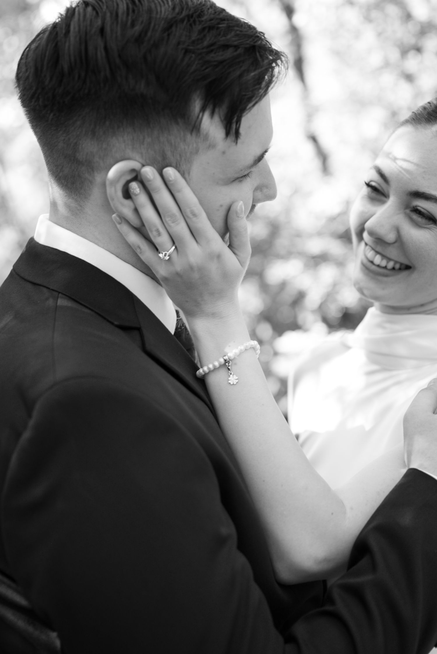 Black and white wedding photo of smiling bride touching grooms face, wearing pearl bracelet and ring outdoors.