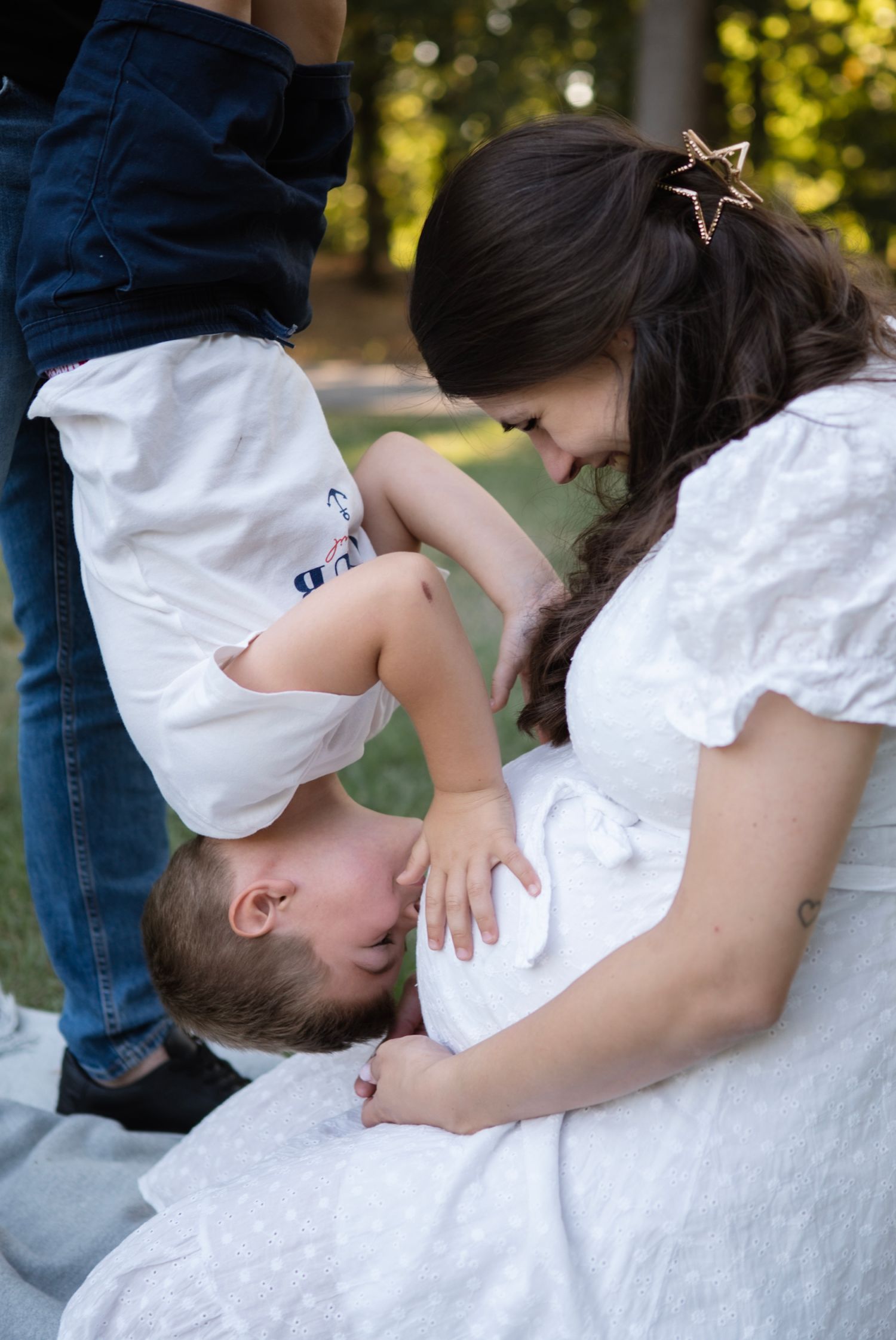 A woman in a white dress smiles at a newborn baby on her lap while a young child leans over to kiss the infant outdoors.