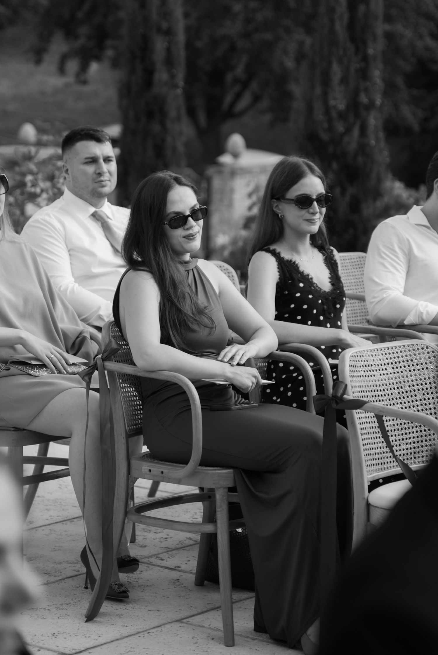 Black and white photo of three guests in sunglasses seated at an outdoor event, two women and a man in formal attire.