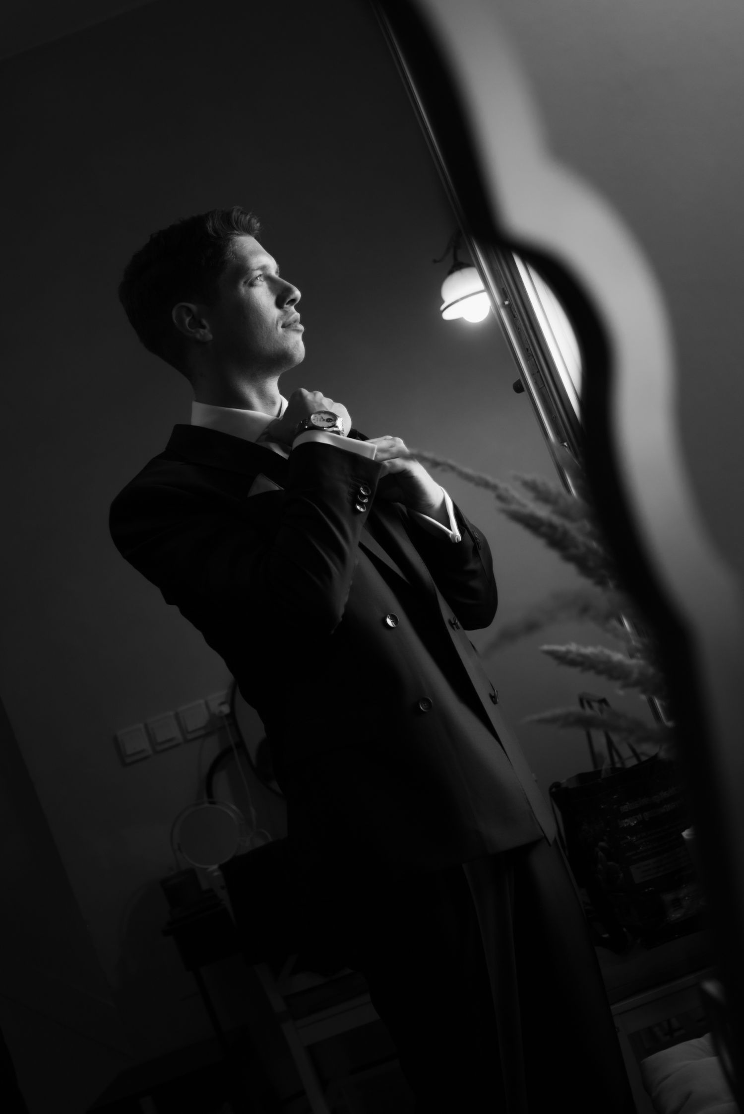 Black and white photo of a groom in a tuxedo adjusting his bow tie, lit by a warm light in an elegant room.