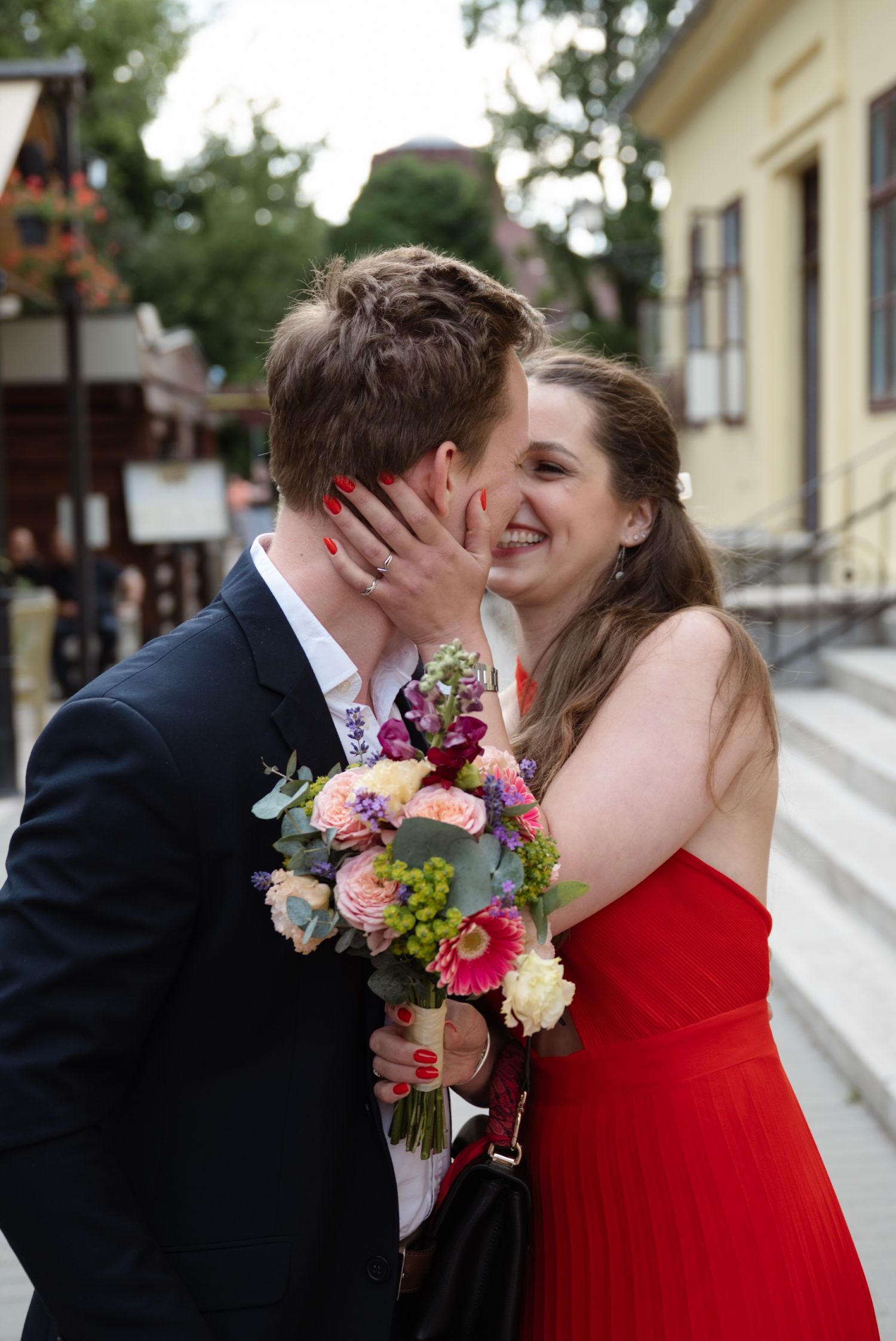Couple sharing a kiss outdoors, woman in red dress holding colorful bouquet, man in navy suit, smiling joyfully.