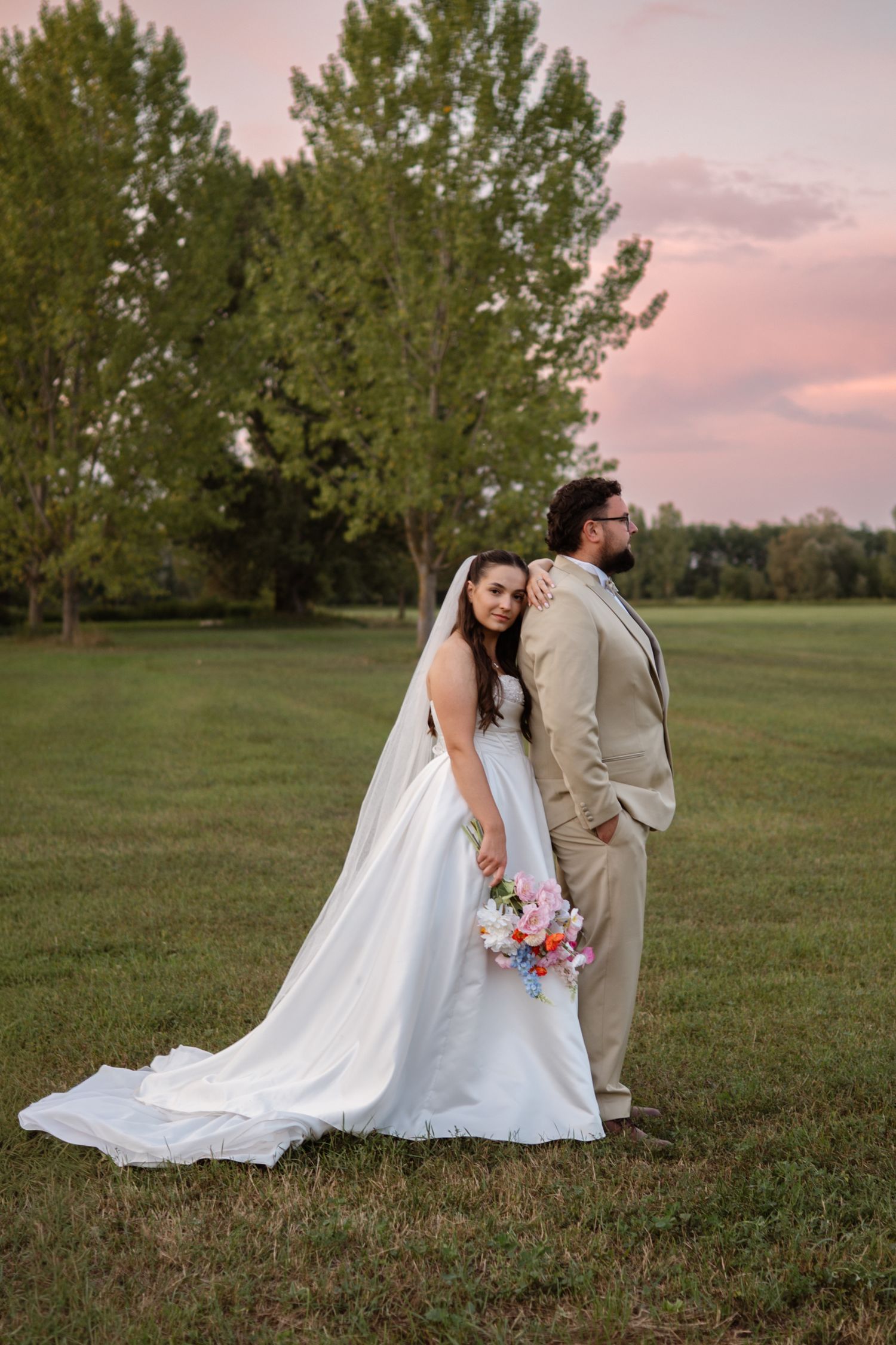 A couple in wedding attire sits together on grass during sunset with a pink sky backdrop.