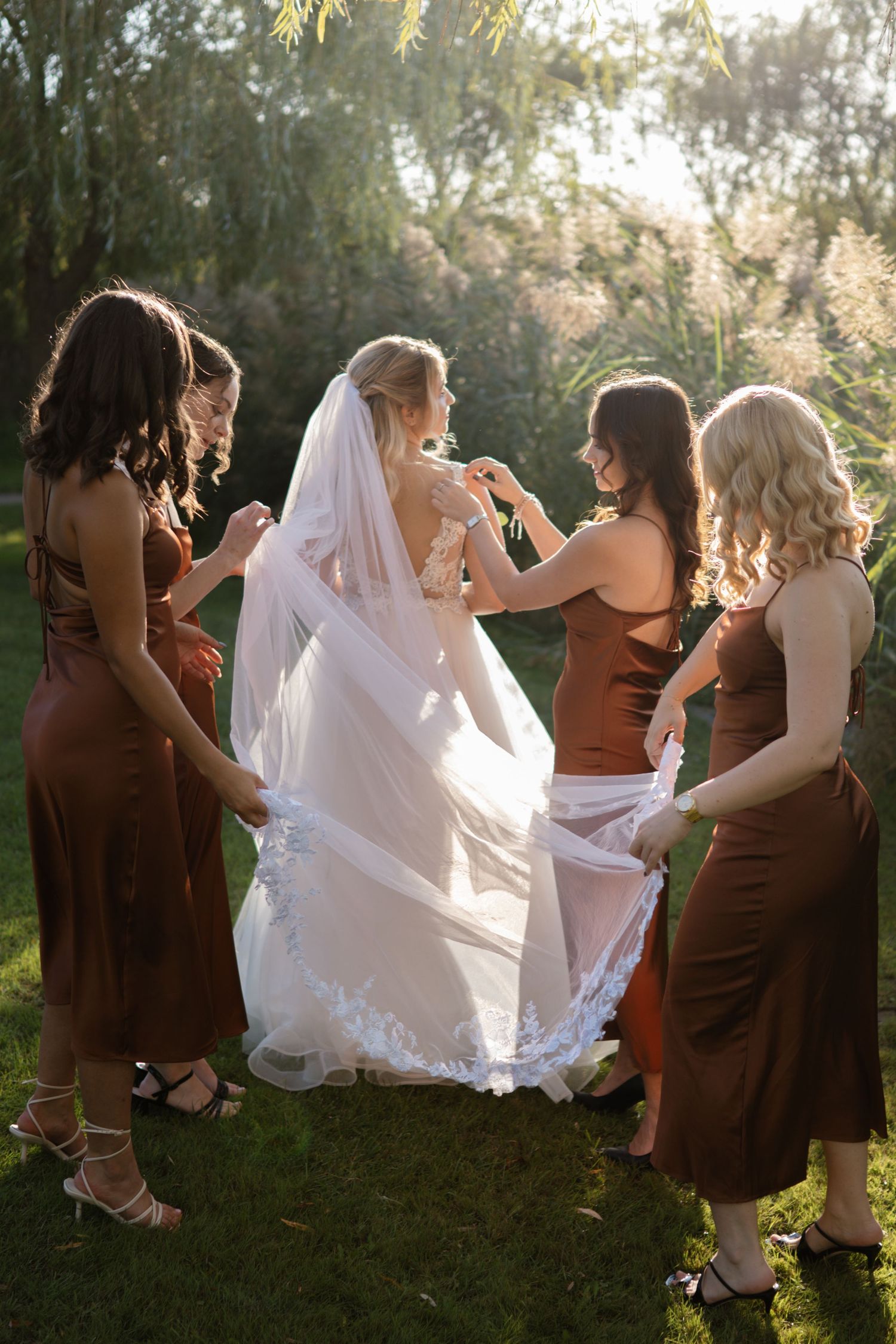 Bridesmaids in brown dresses help adjust the bride's white wedding dress train outdoors.