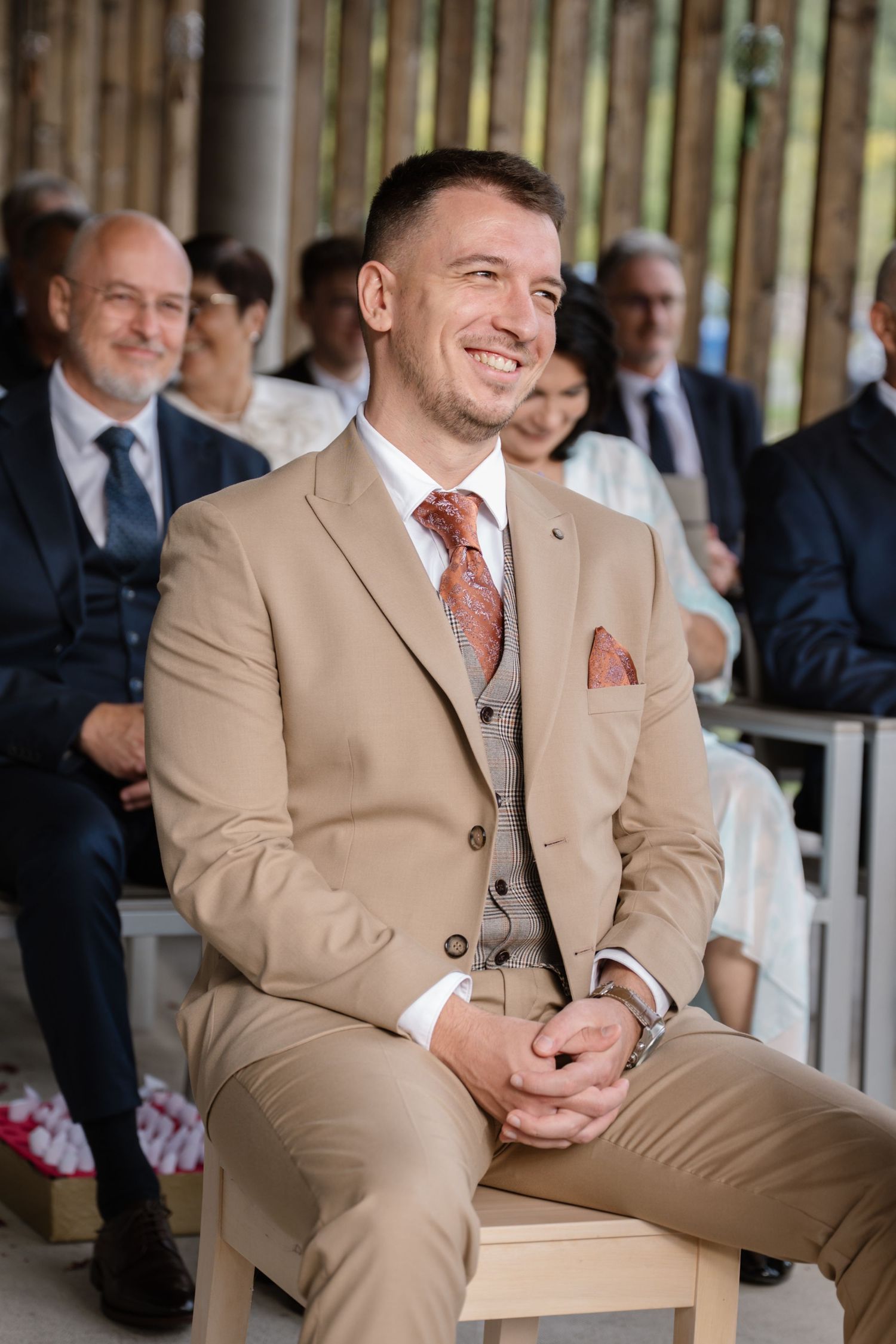 A guest in a beige three-piece suit smiles during an outdoor wedding ceremony.