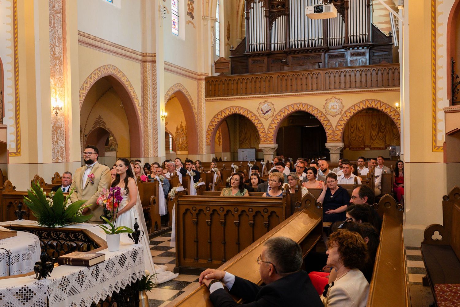 Wedding ceremony taking place in an elegant church with ornate architecture and wooden pews filled with guests.