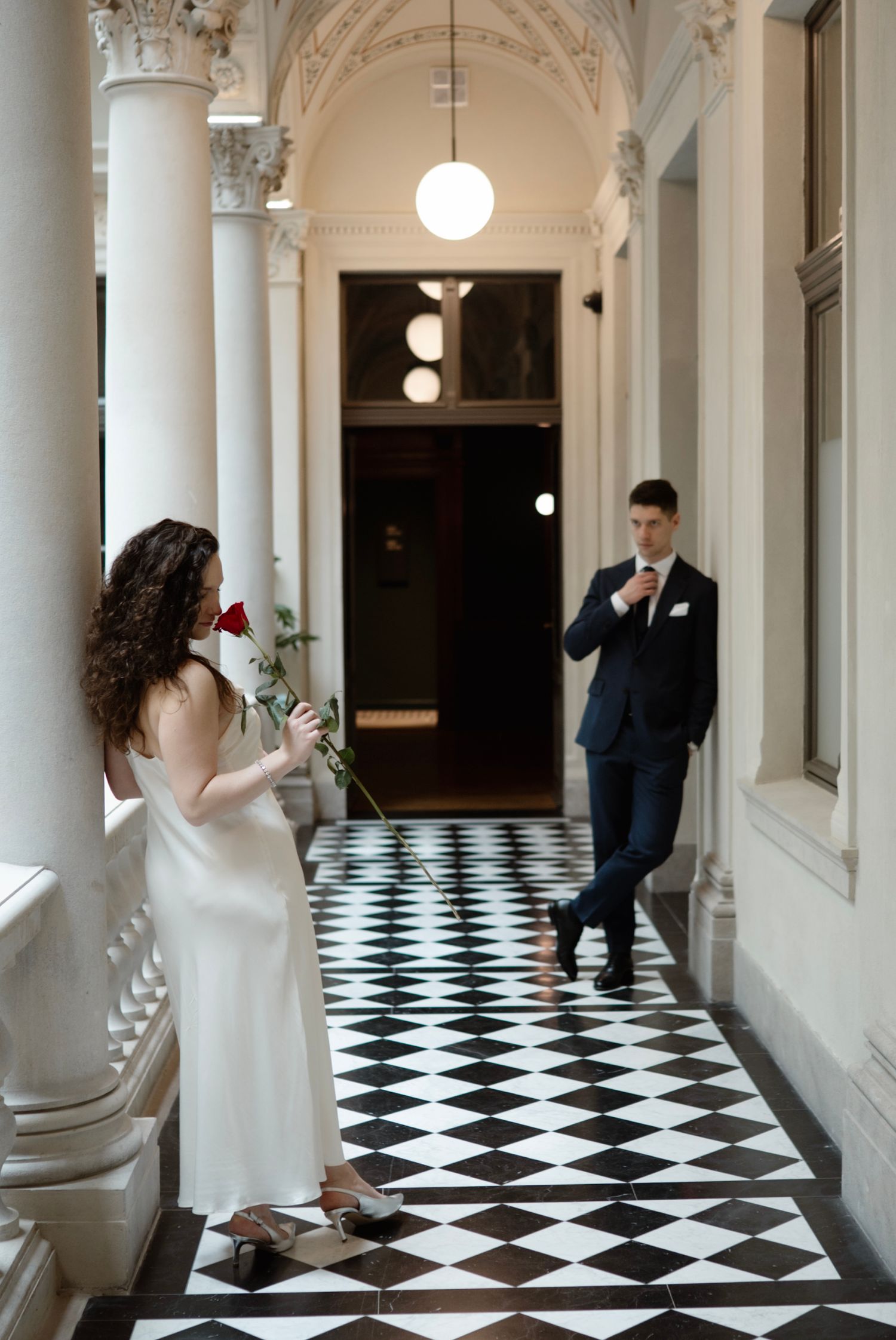 Bride in white slip dress holds red rose while groom in navy suit adjusts tie in elegant checkered hallway.