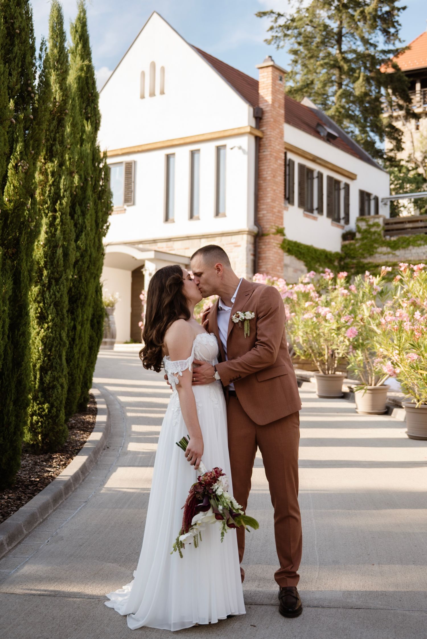 Bride and groom share a kiss outside an elegant white manor, surrounded by cypress trees and blooming flowers.