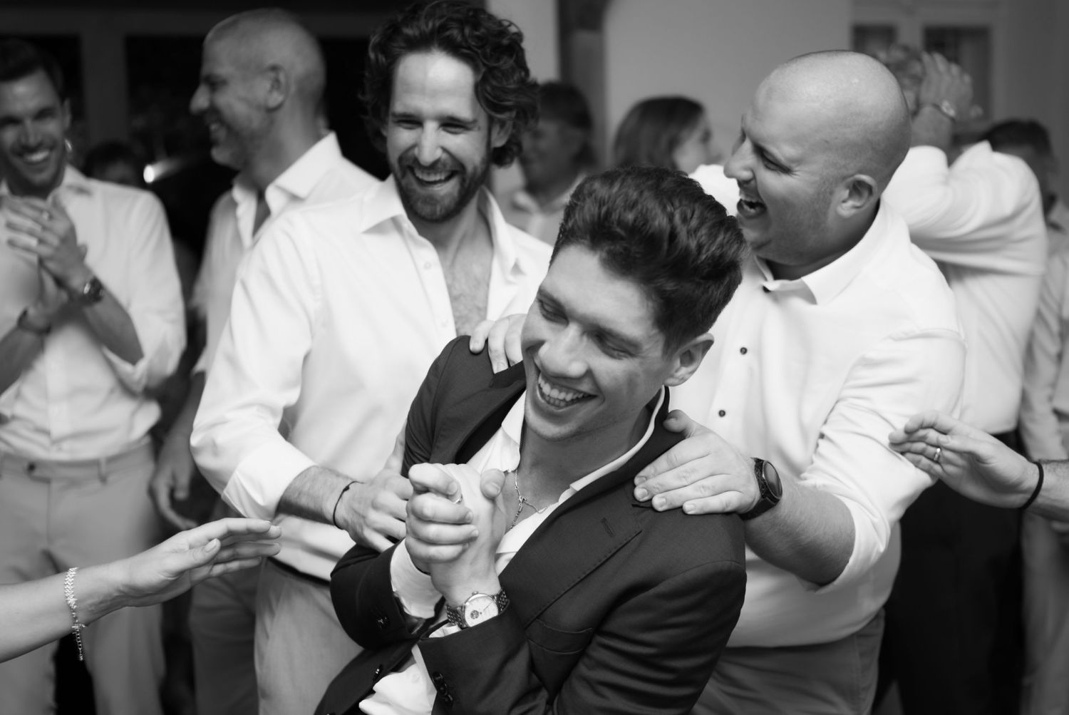 Men celebrating joyfully at a wedding reception, laughing and dancing together in black and white photo.