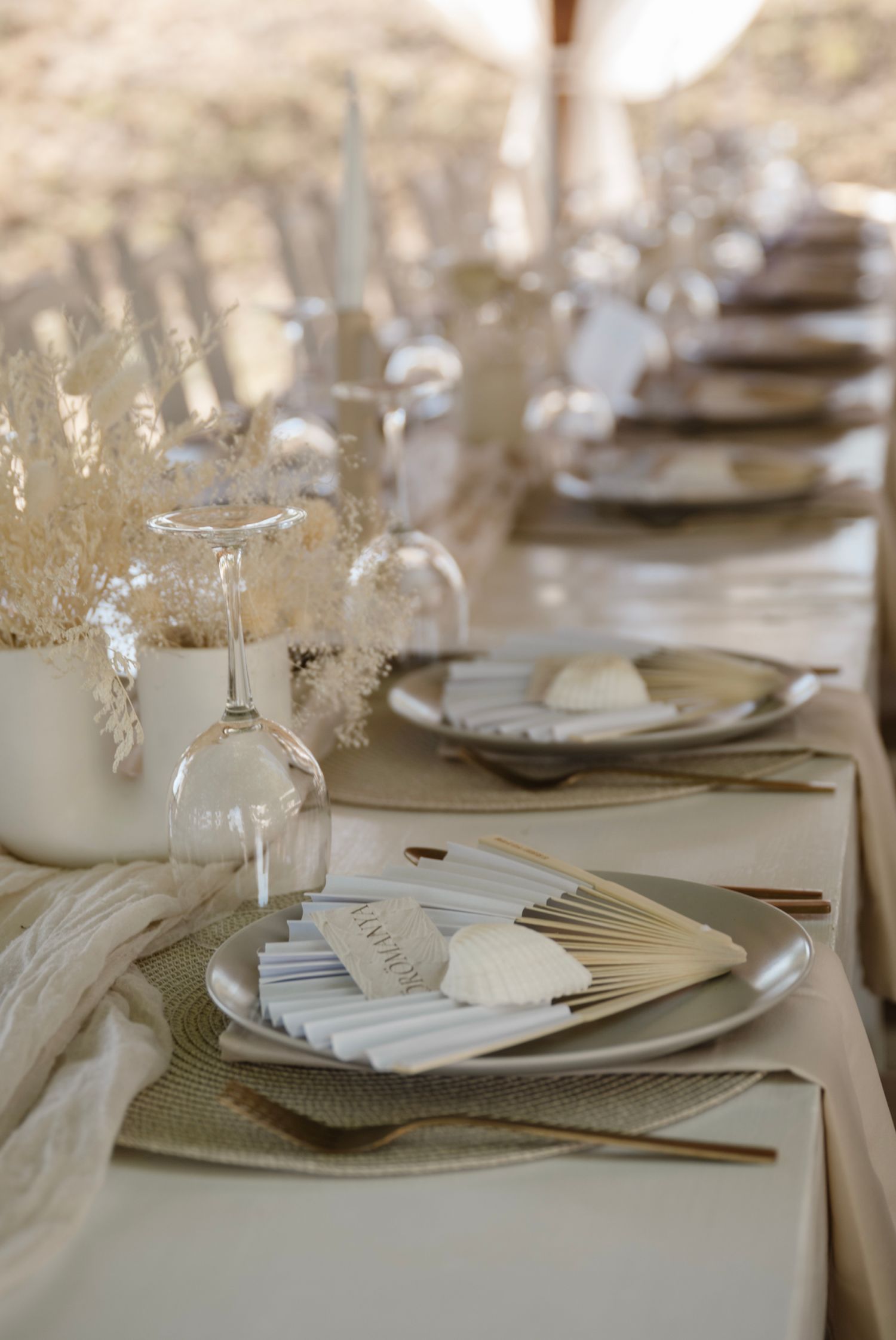 Elegant wedding table setting with neutral tones, gold cutlery, pampas grass, and decorative fans on silver plates.