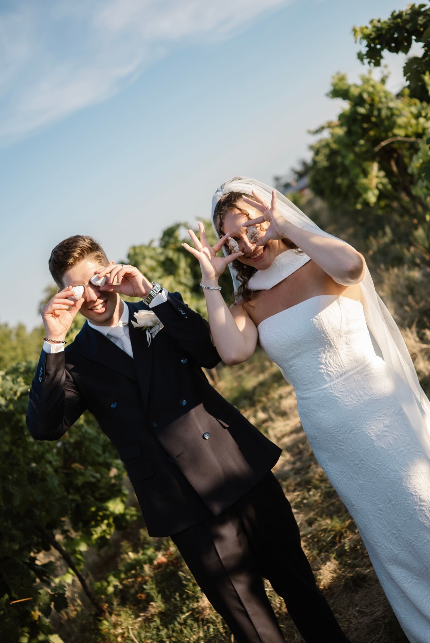Joyful bride and groom making heart and peace signs at vineyard wedding, bride in white gown with veil, groom in dark suit.