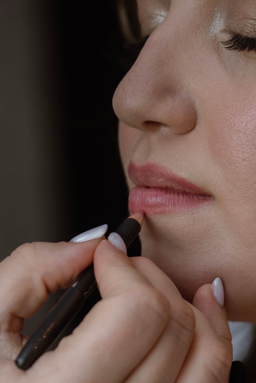 A makeup artist applies lip liner pencil to a woman's lips with white manicured nails visible.