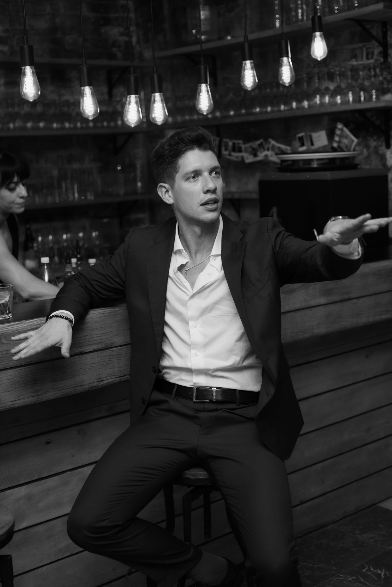 Man in suit sitting at bar gesturing with hands, black and white photo with Edison bulb lighting.