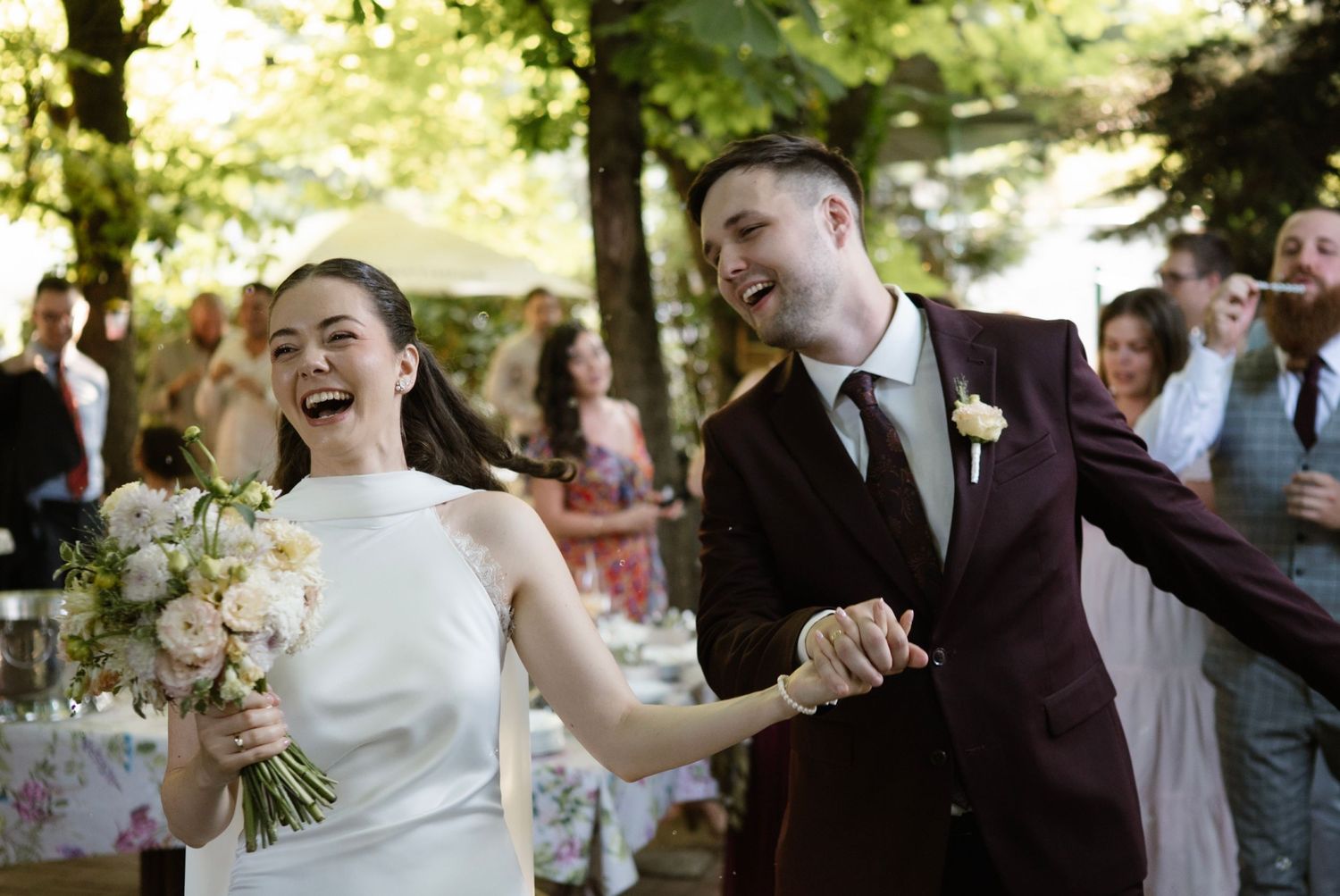 Joyful newlywed couple holding hands and laughing at outdoor wedding reception, bride holding floral bouquet.