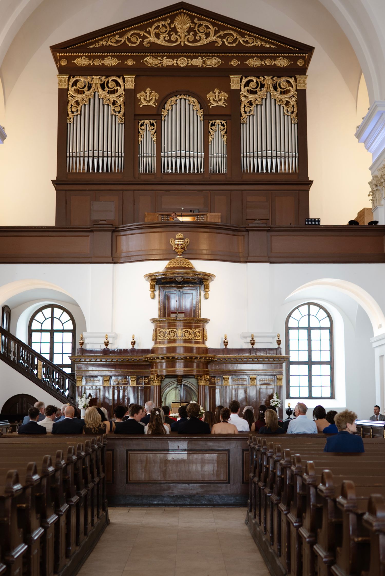 Church interior with ornate pipe organ, wooden pews filled with congregation, and decorative baroque pulpit during a service.