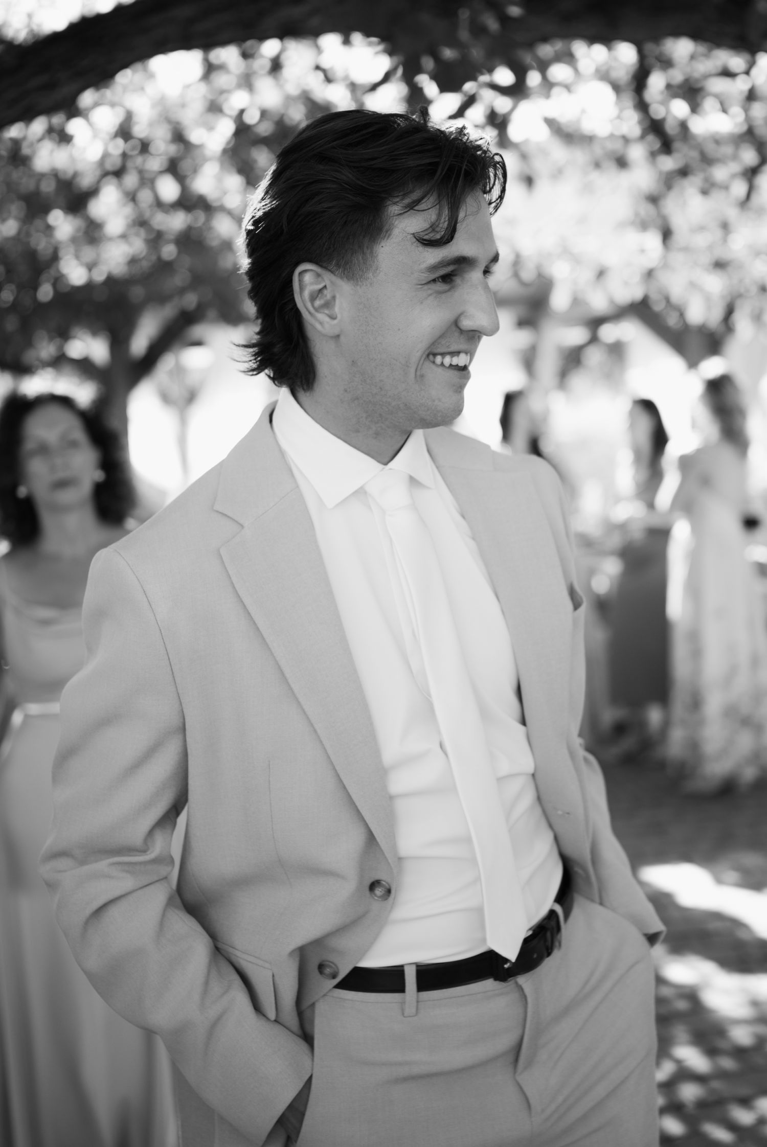 Smiling groom in light gray suit and tie at outdoor wedding ceremony, black and white portrait photography.