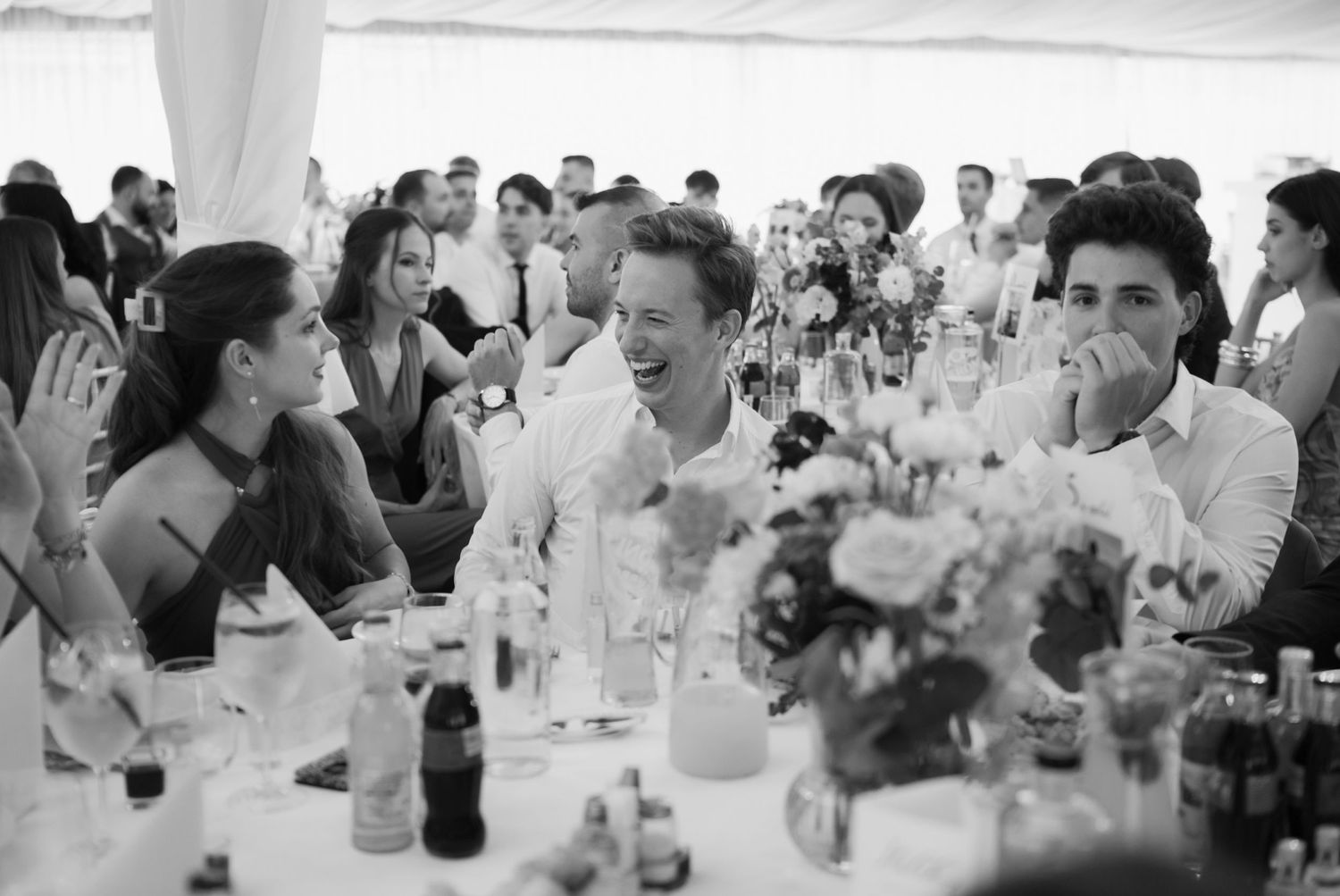Guests laughing joyfully at a wedding reception table in a marquee, captured in black and white.