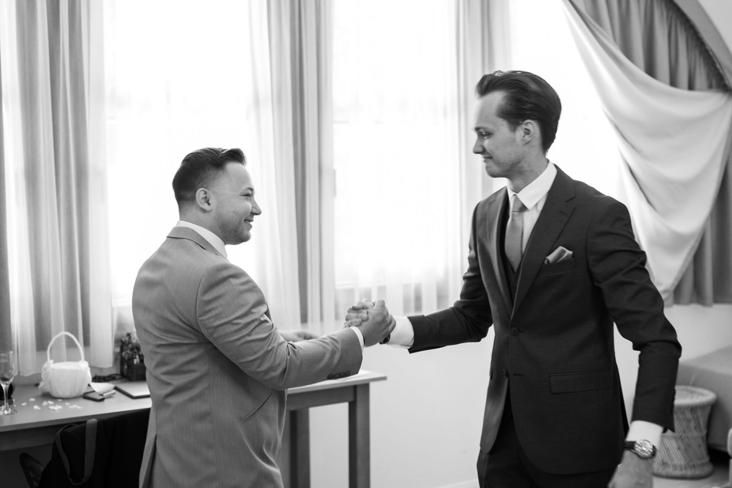 Two men in suits shaking hands warmly in an indoor setting with curtains in the background.