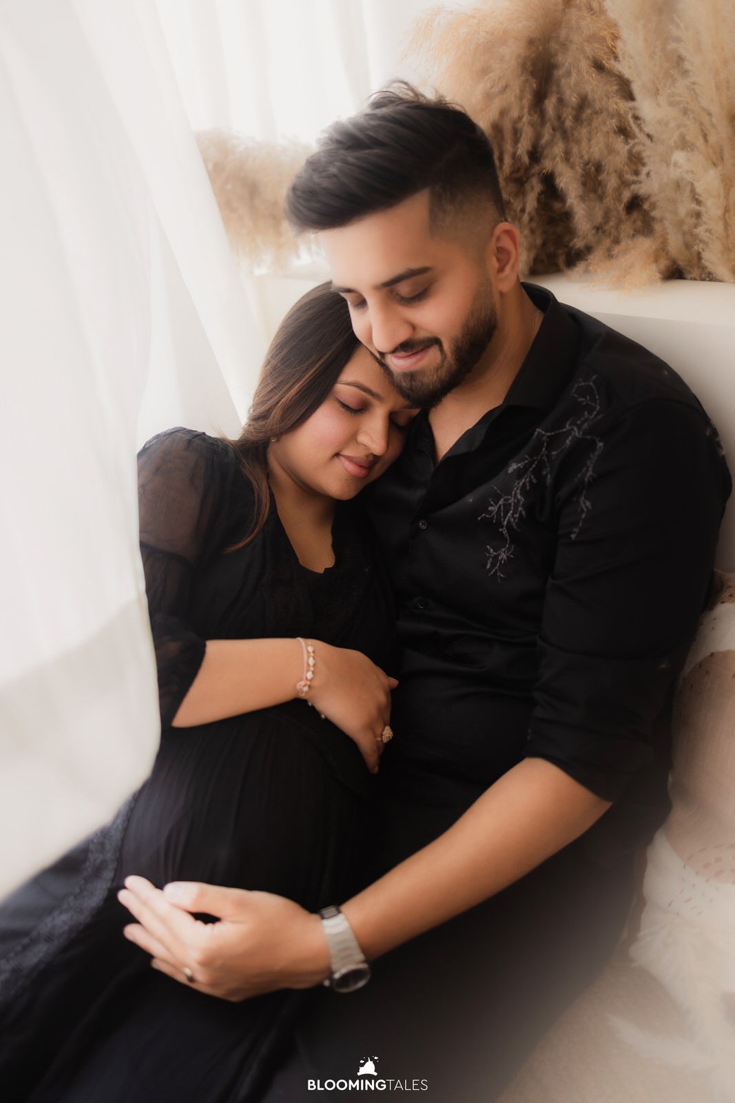 A couple in black outfits share an intimate moment during a maternity photoshoot with soft lighting.
