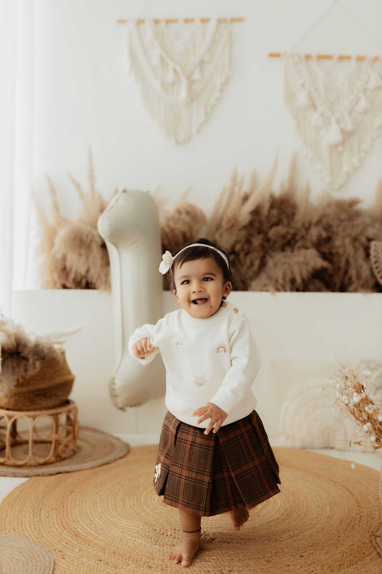 A toddler in a white sweater and plaid skirt dances joyfully on a round rug surrounded by boho decor.