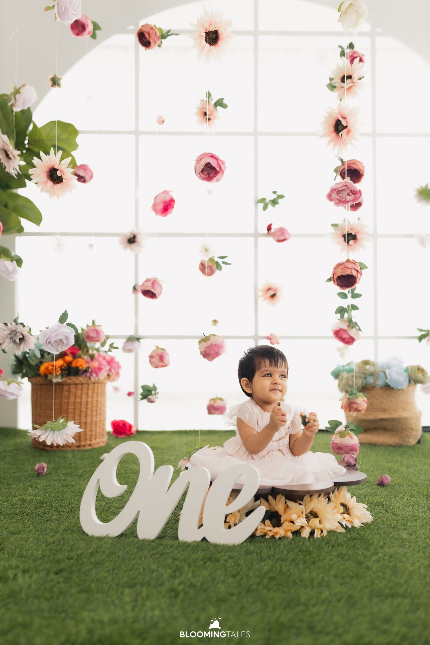 First birthday photoshoot with floral decorations and 'one' text prop on artificial grass.