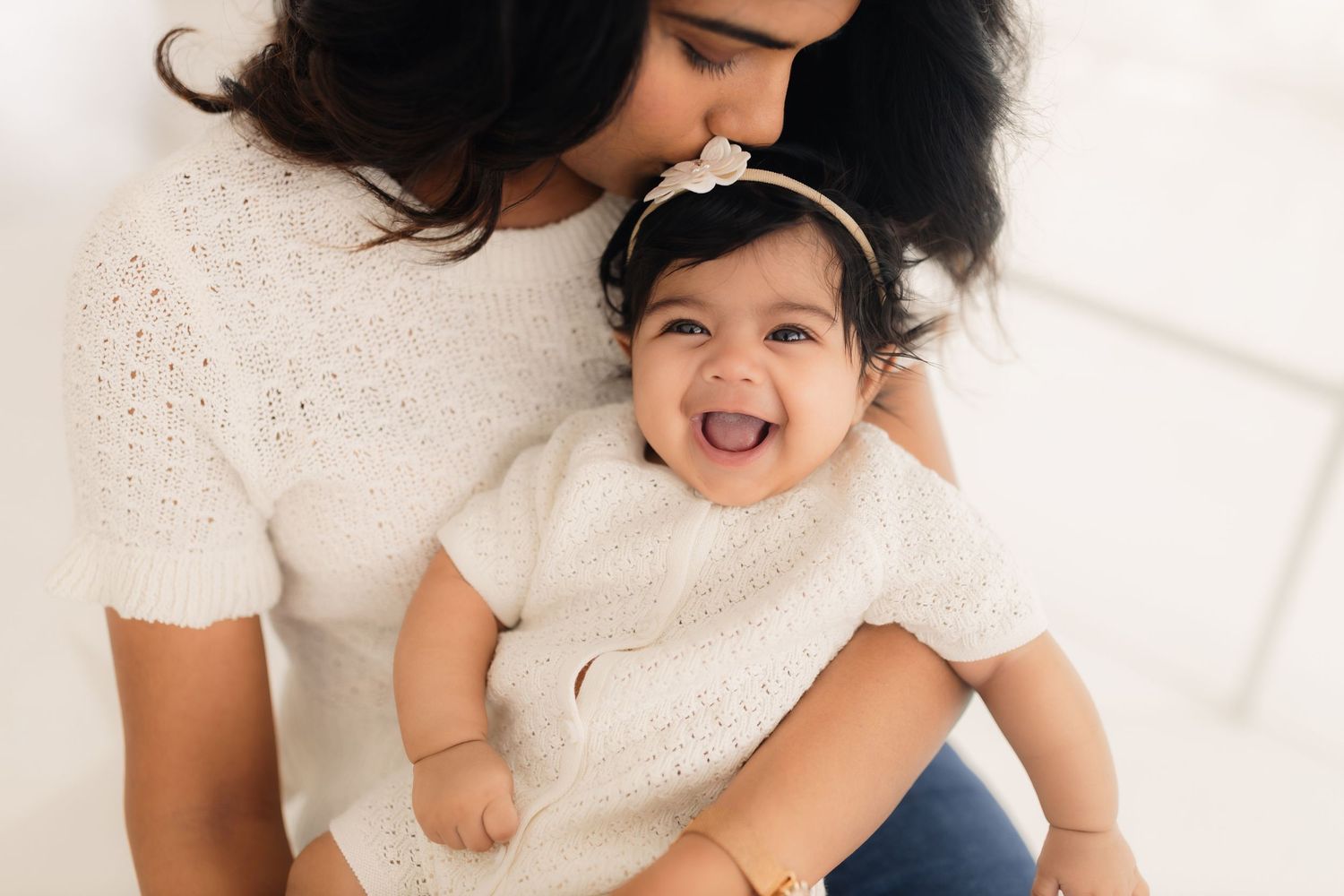 Mother and baby share joyful laughter while sitting together on white bedding.