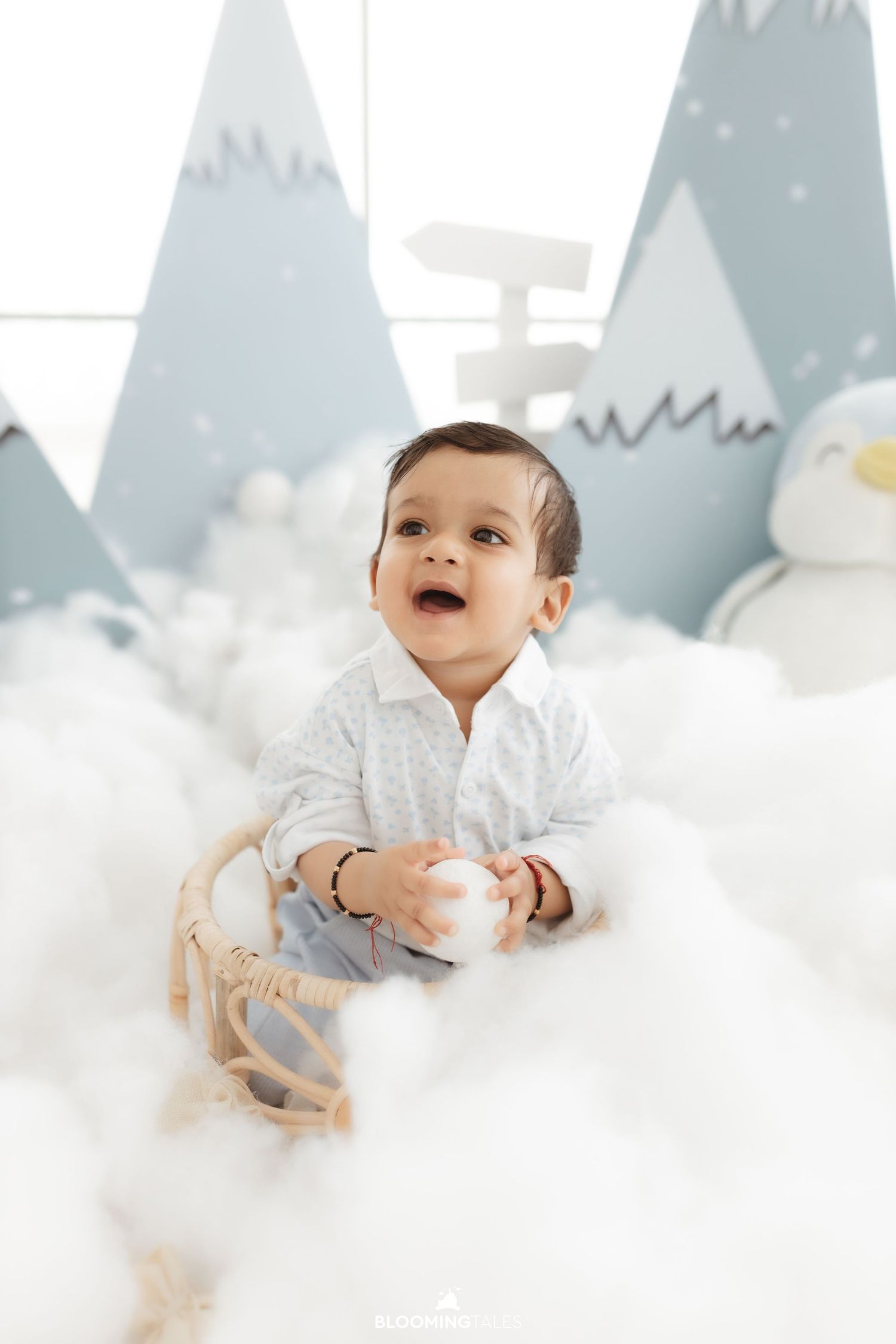 Baby in white outfit sits among cotton cloud decorations with painted mountain backdrop.