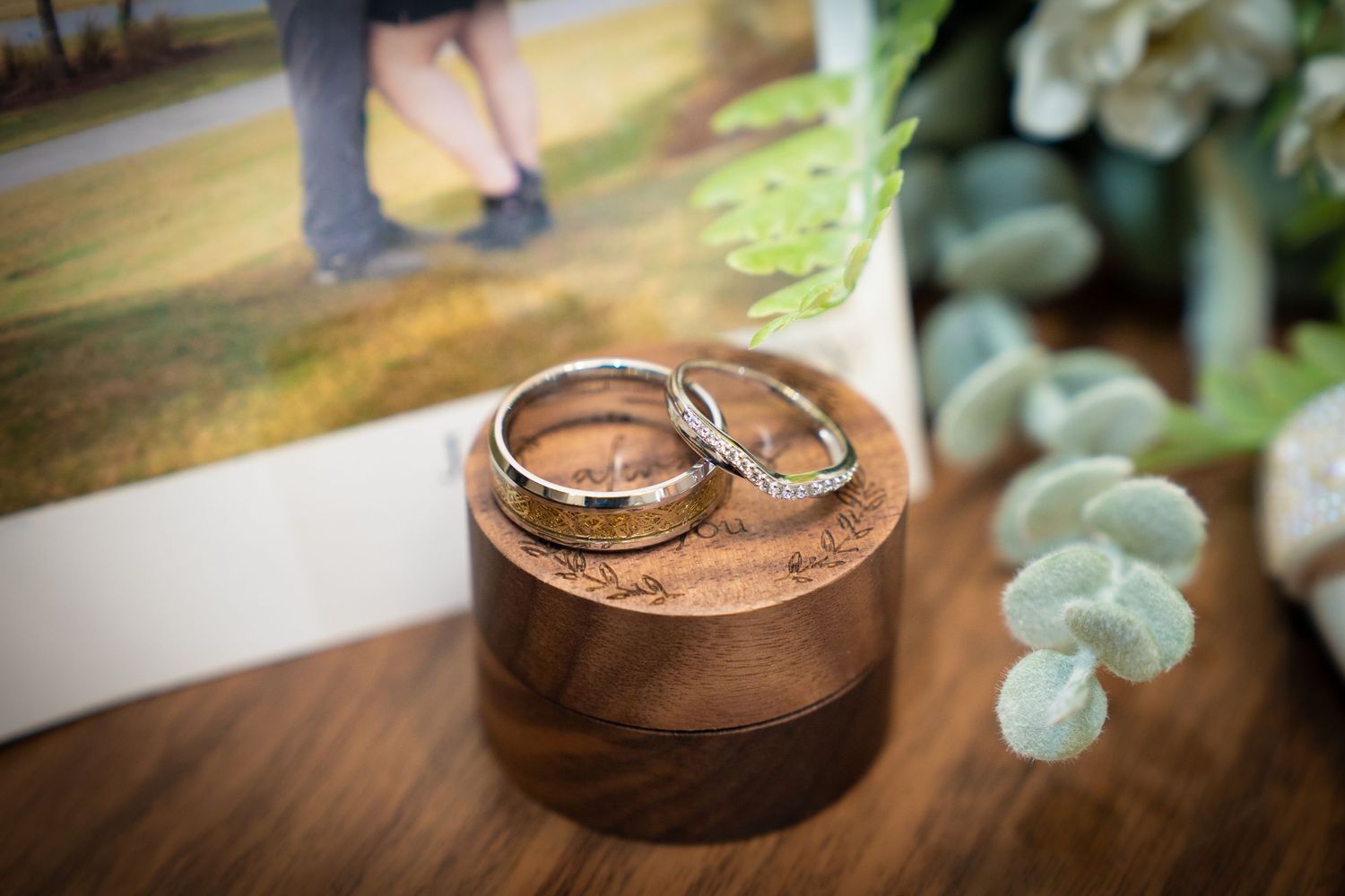 Two wedding rings nestled in a rustic wooden bowl with soft green eucalyptus leaves in the background.