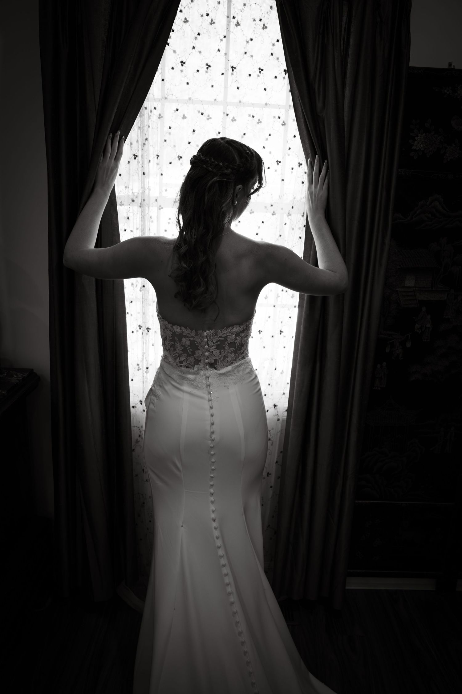 Black and white photo of a bride in a fitted wedding gown standing at a window with curtains, viewed from behind.