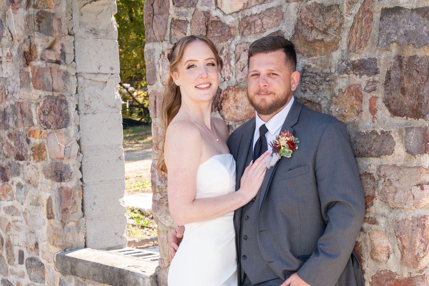 Bride and groom posing together against a rustic stone wall, smiling on their wedding day.