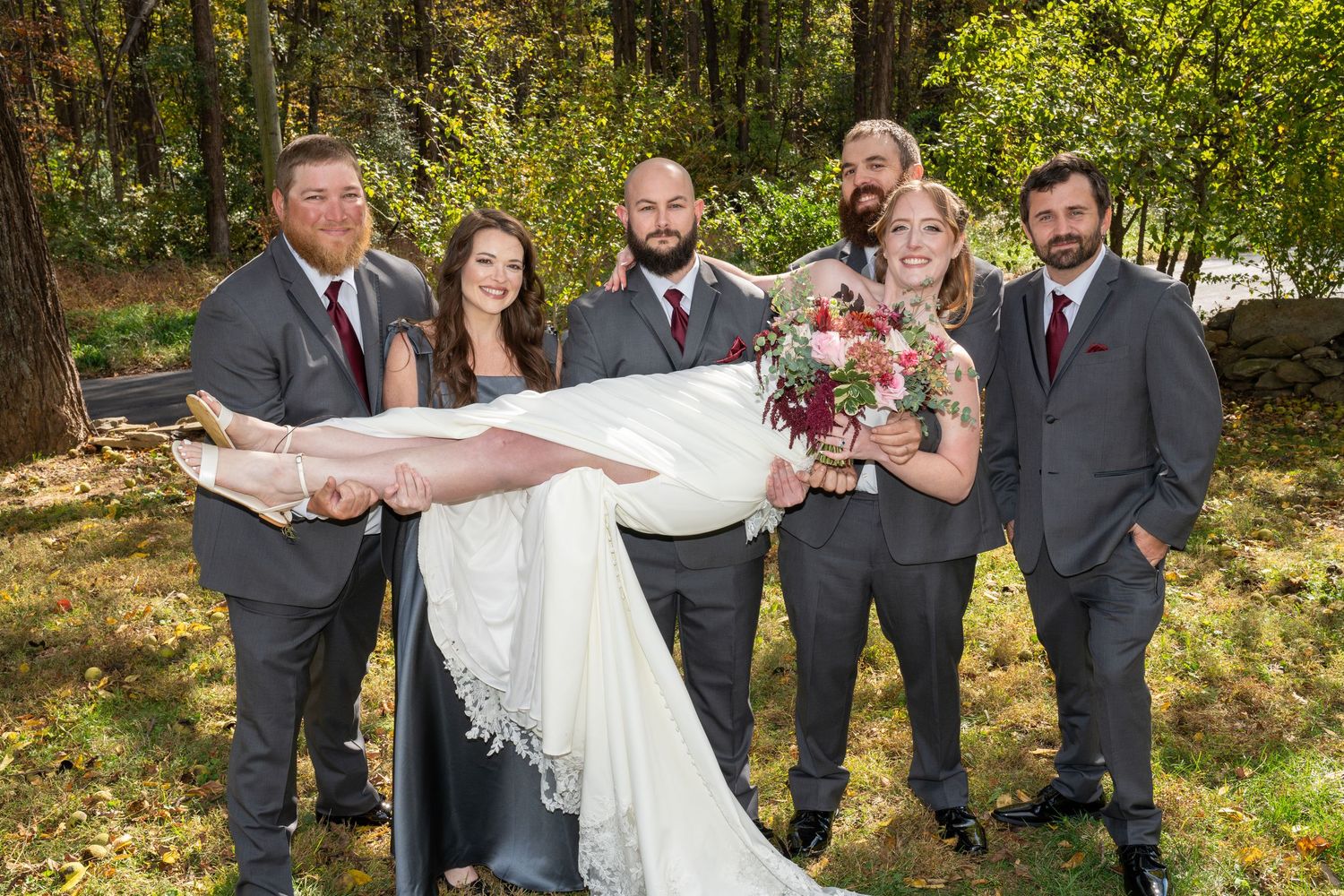 Groomsmen in gray suits carry smiling bride in white wedding dress outdoors among autumn trees.