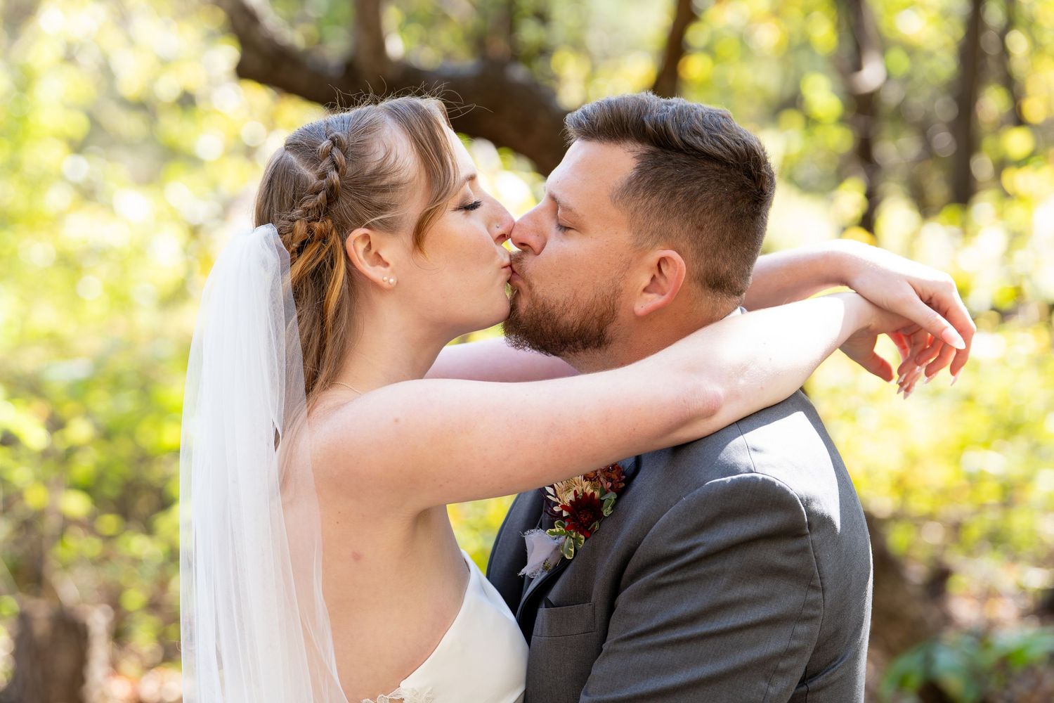 Bride and groom sharing a romantic kiss outdoors, bride wearing veil with braided updo, groom in gray suit.