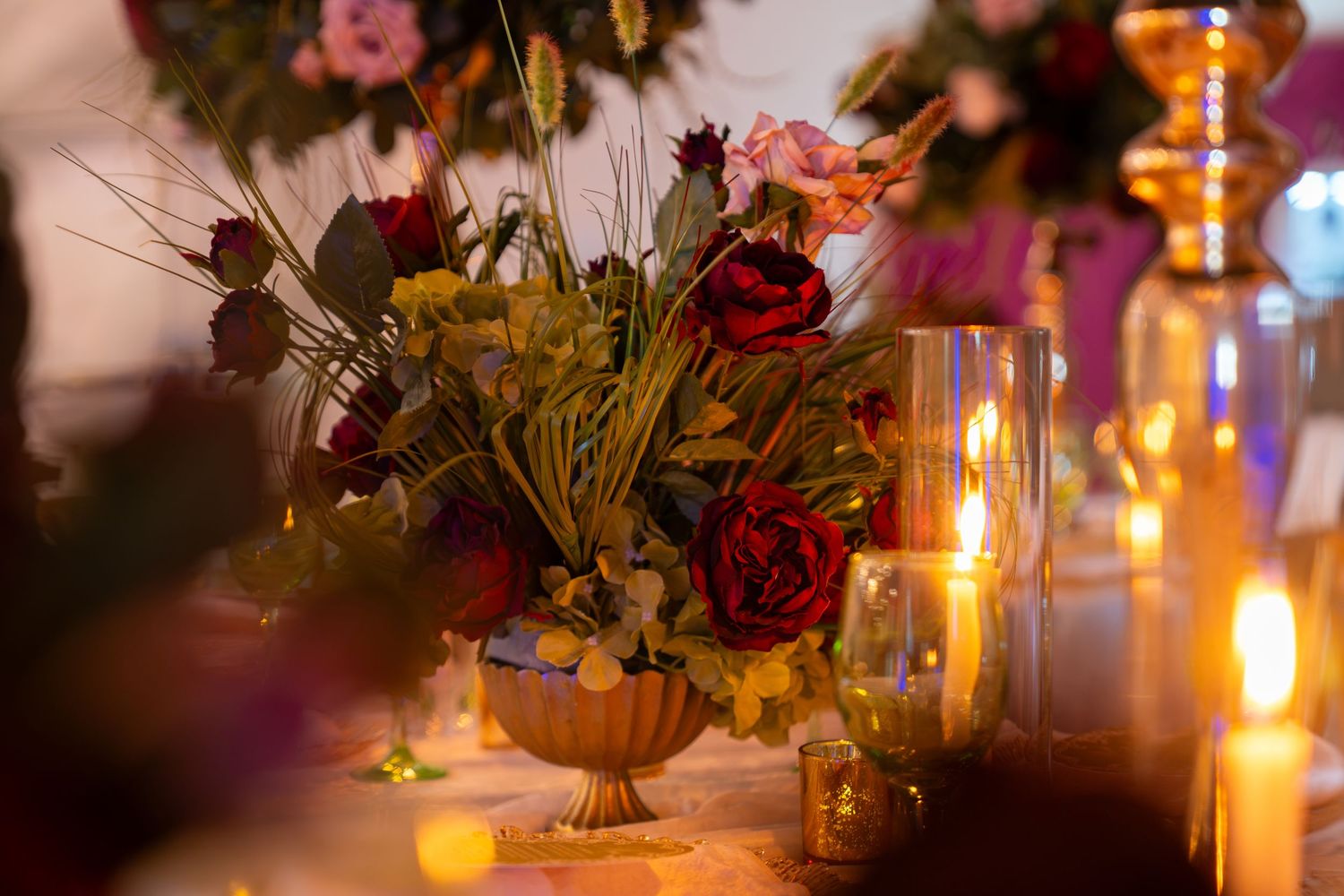 Elegant wedding table centerpiece with red roses in gold vase, surrounded by glowing candles in warm amber light.