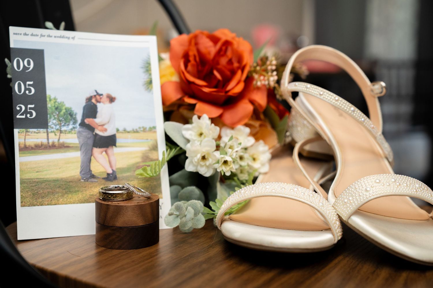 Wedding details arranged on dark wood surface including heeled sandals, orange roses, and a framed photo.