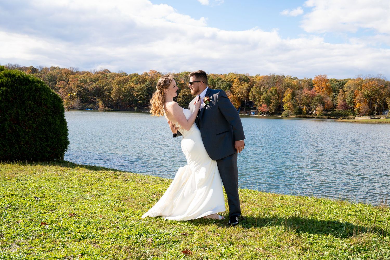A couple in formal attire poses on a grassy shore with autumn trees and a lake in the background.
