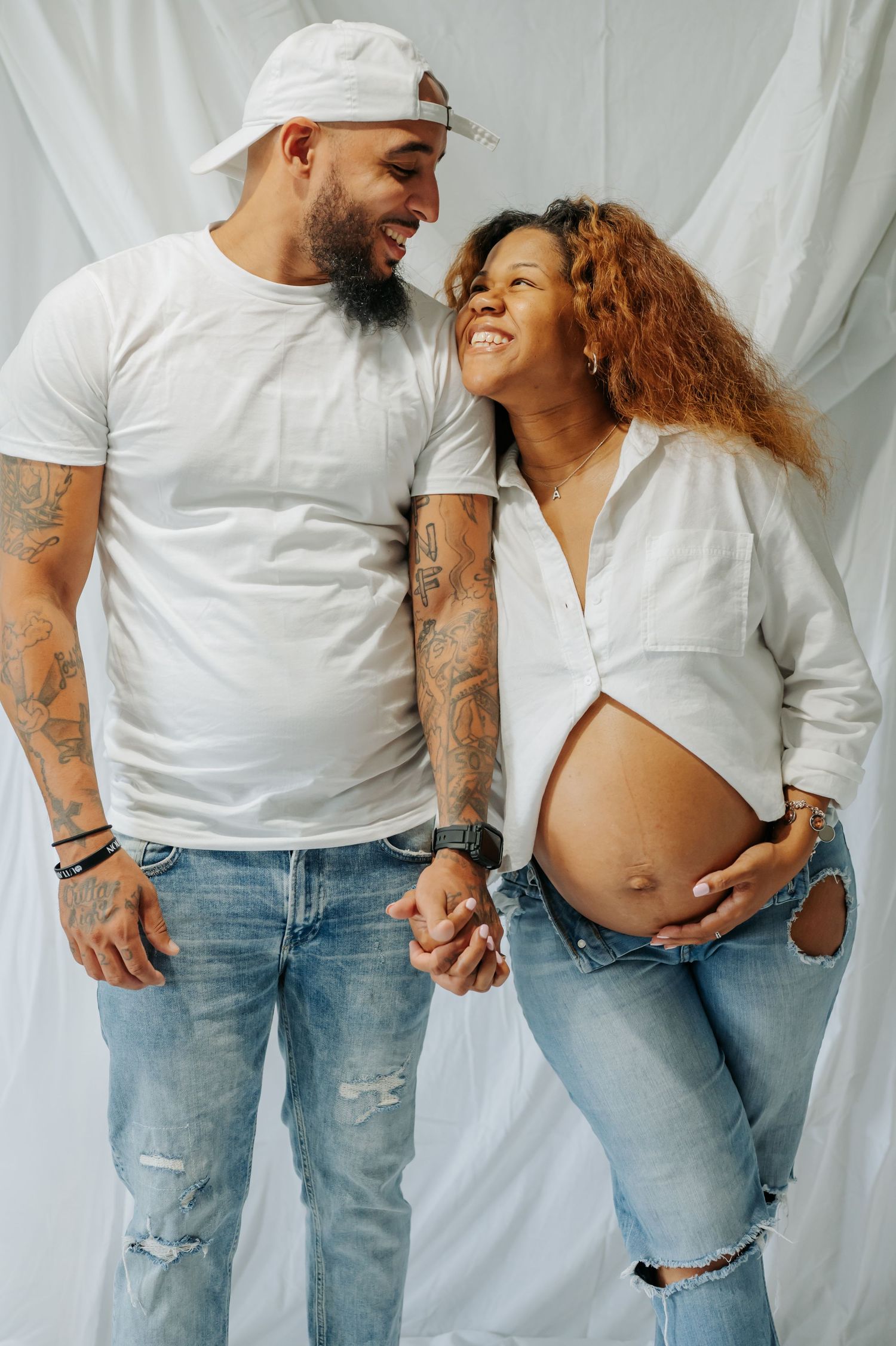 Casual couple in matching white tops and distressed jeans posing playfully against white background.