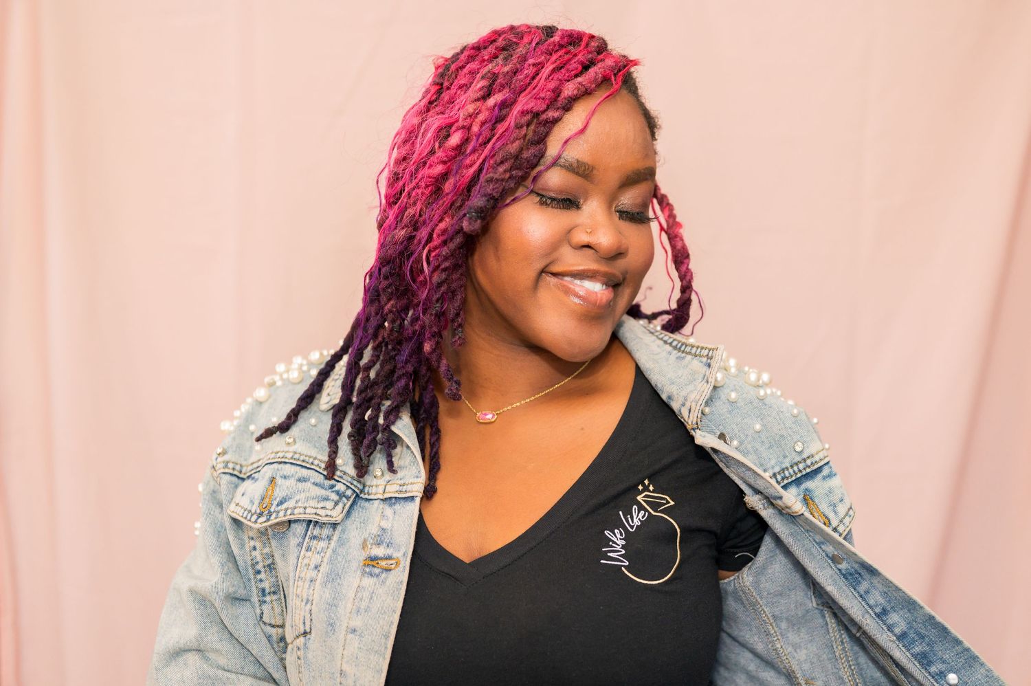 A person with bright pink braids smiles warmly while wearing a denim jacket and black top against a pink backdrop.
