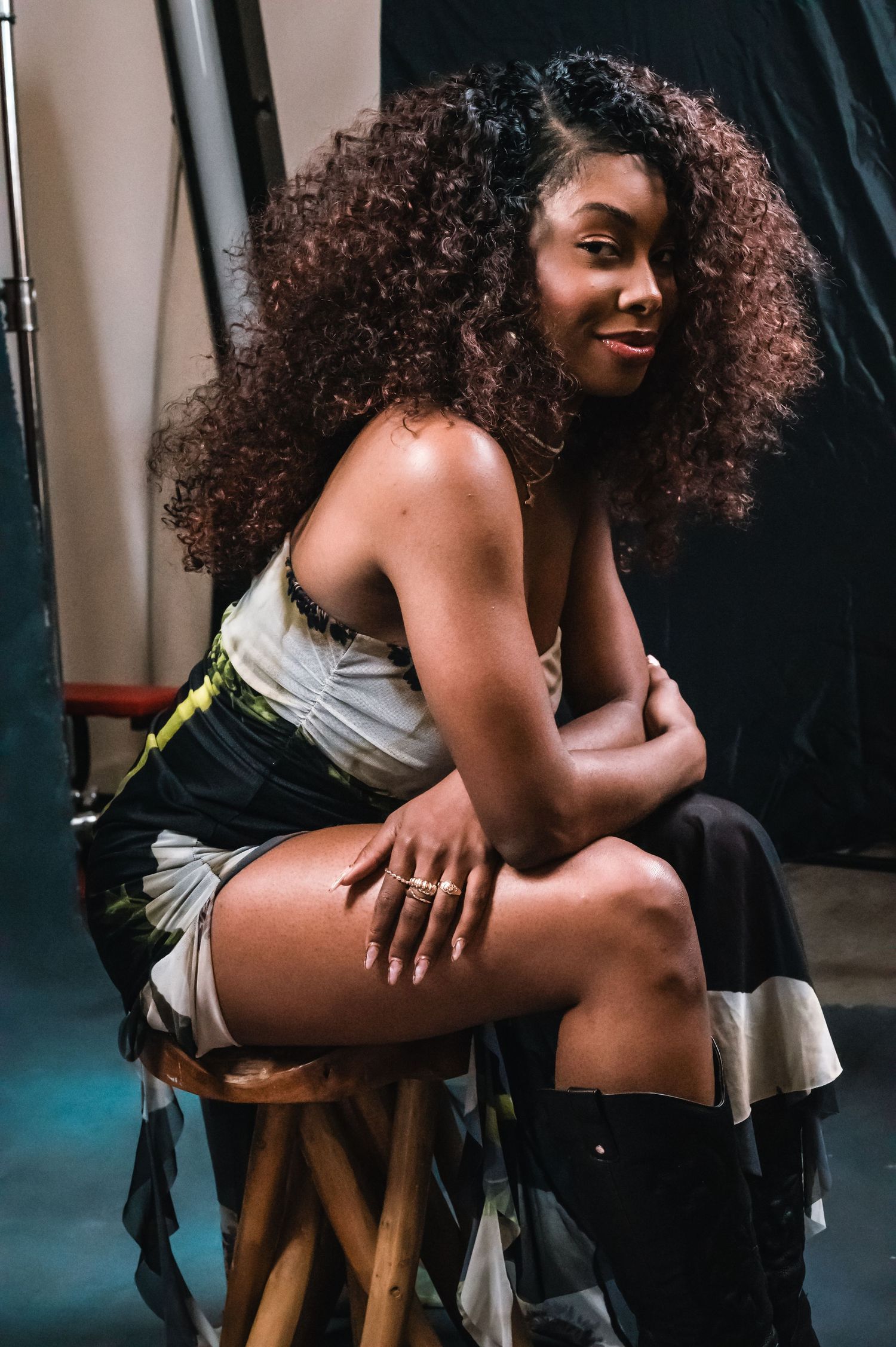 Young woman with curly hair poses on a wooden stool in a studio, wearing a colorful mini skirt and black boots.