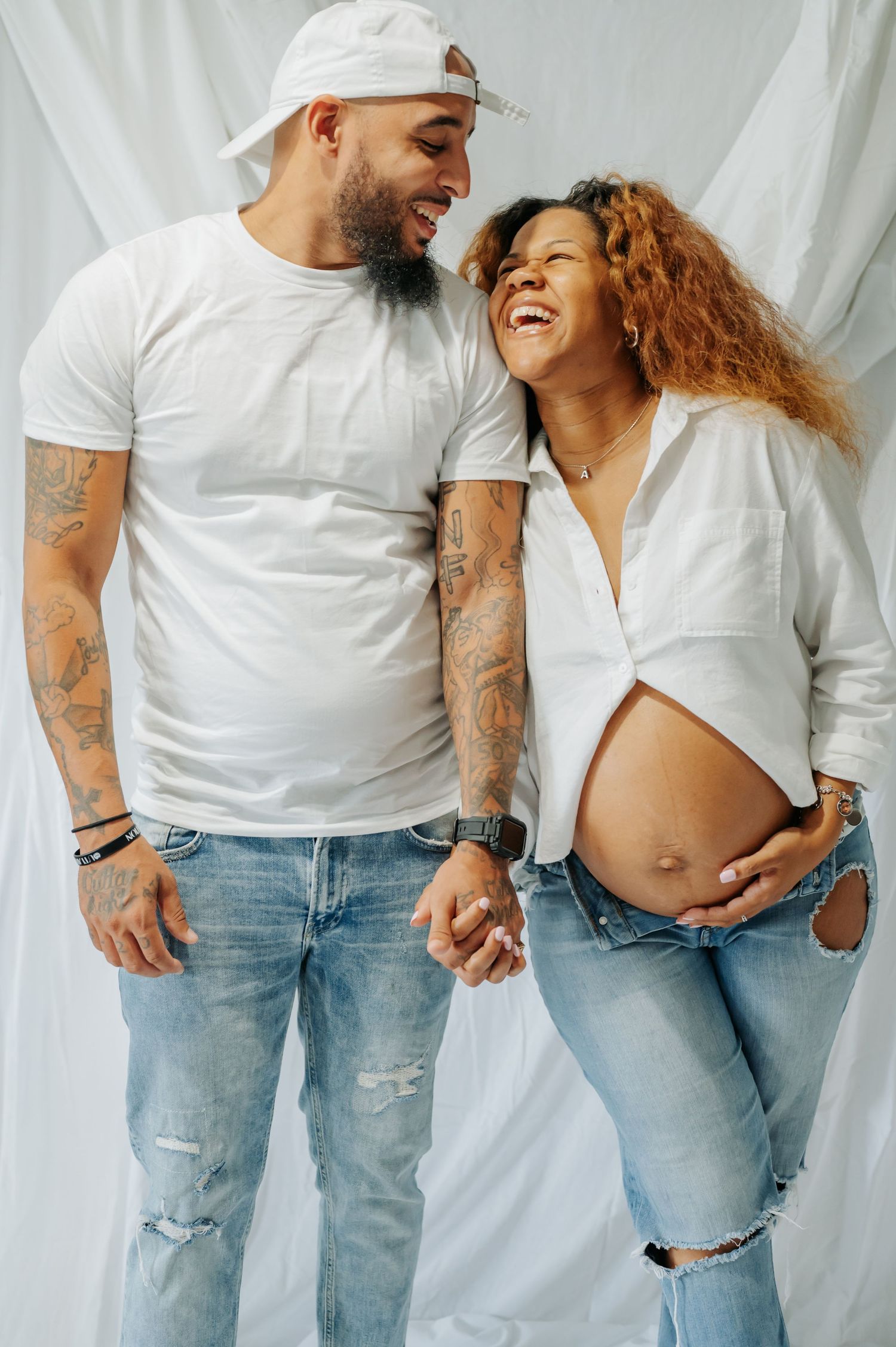A couple in matching white tops and ripped jeans share a joyful moment while showing off her pregnant belly against white backdrop.