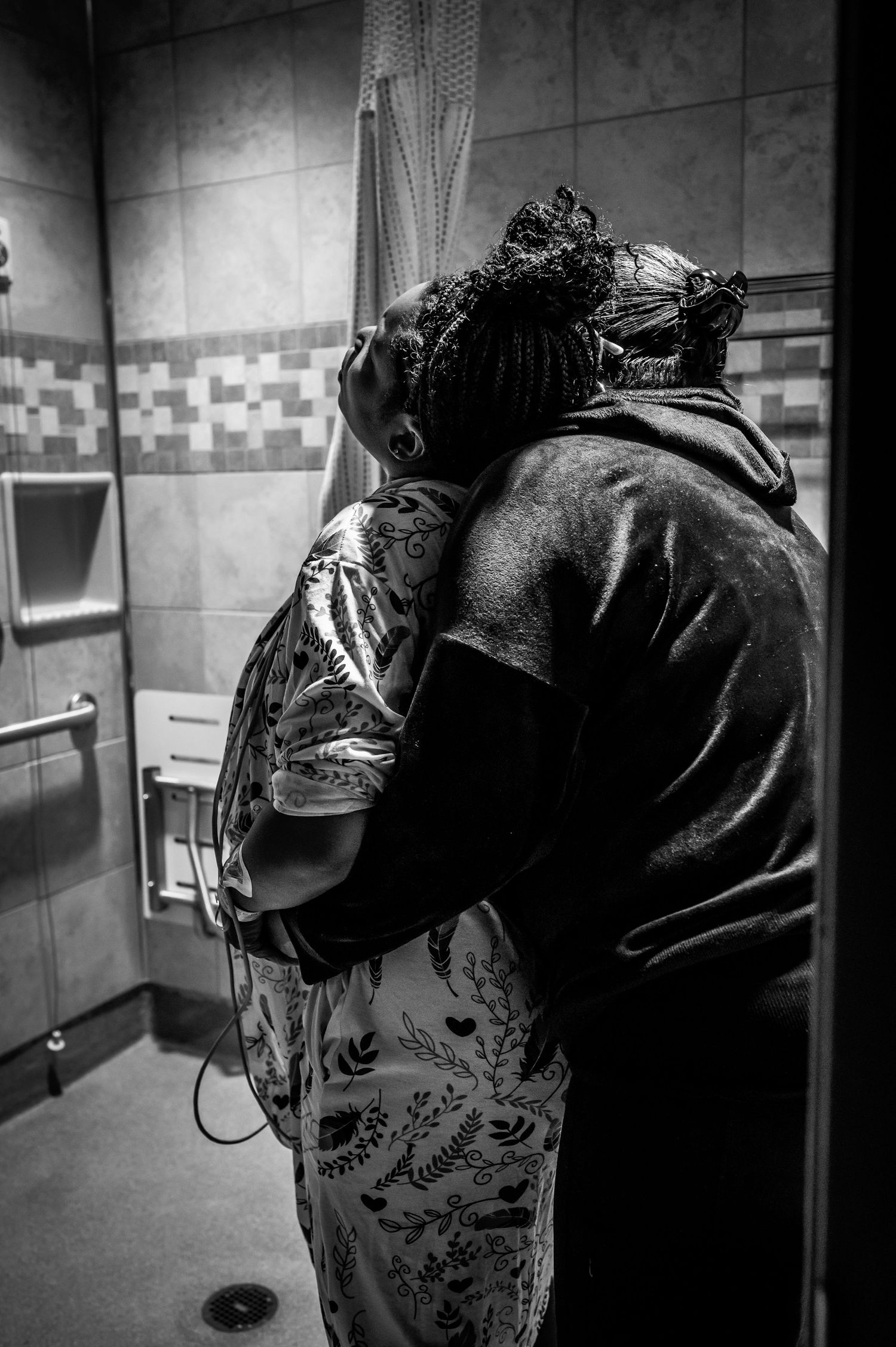 Black and white photo of person with curly hair hugging another in a bathroom with tiled walls.
