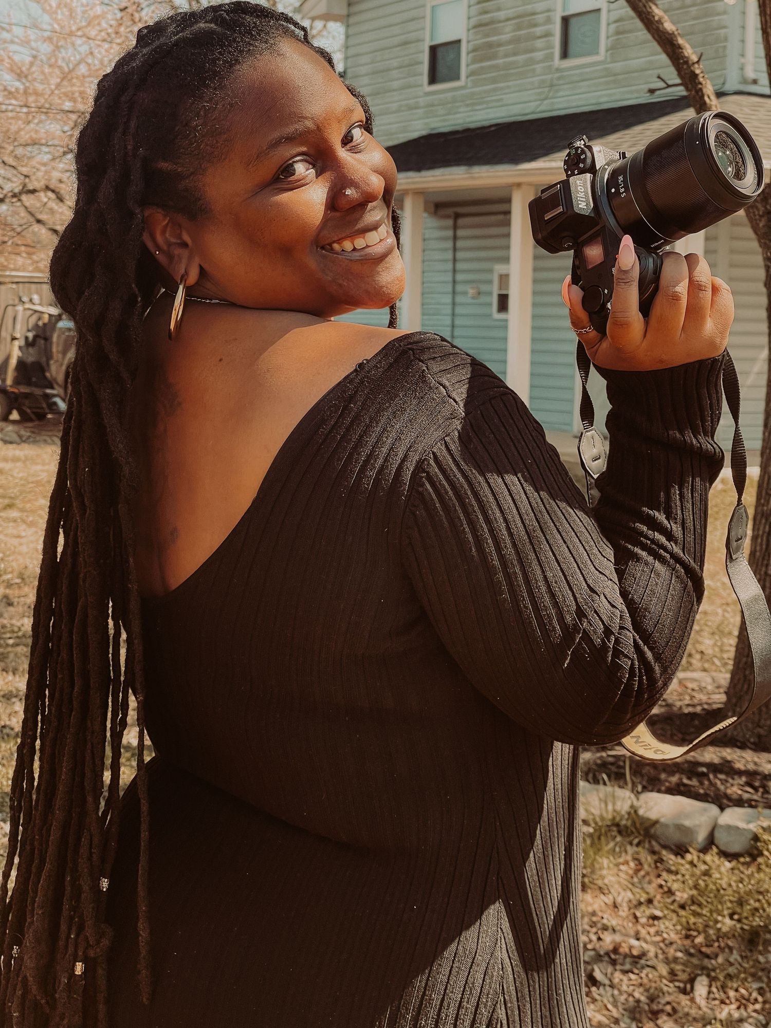 Professional photographer in a black sweater holding a camera outdoors in warm sunlight.
